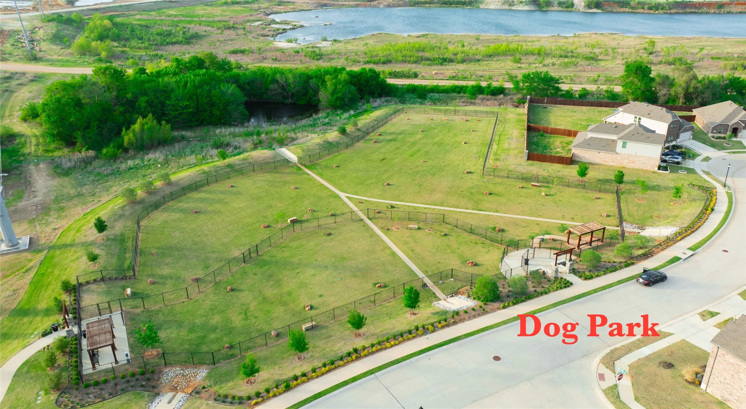 Expansive fenced community dog park featuring multiple grassy areas, paved walking paths, and two shaded seating structures