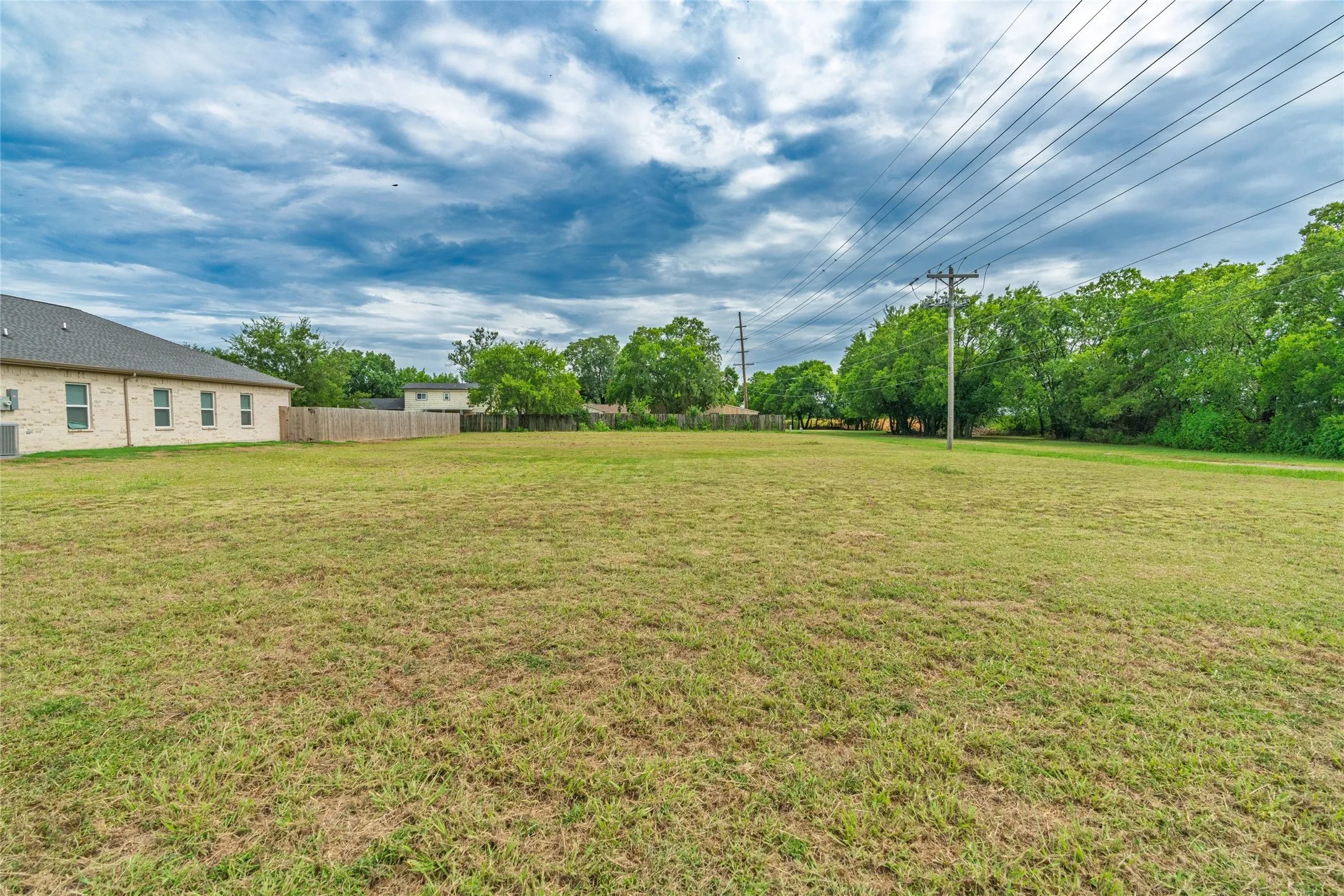 Expansive grass lot featuring a partial wooden fence, mature trees bordering the property, and power lines