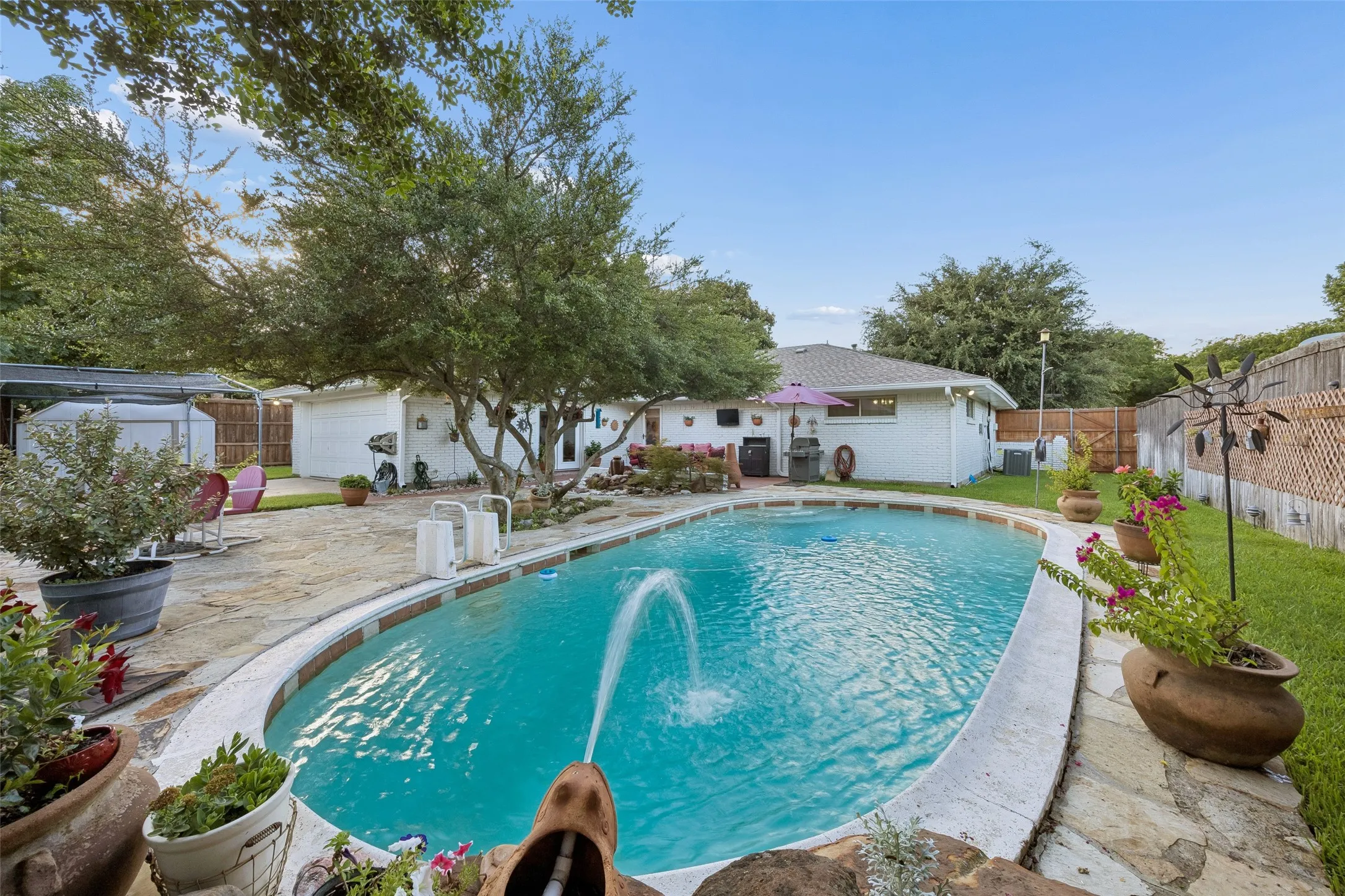 Backyard oasis featuring an oval swimming pool with water features, extensive stone patio, mature trees, and a white brick exterior