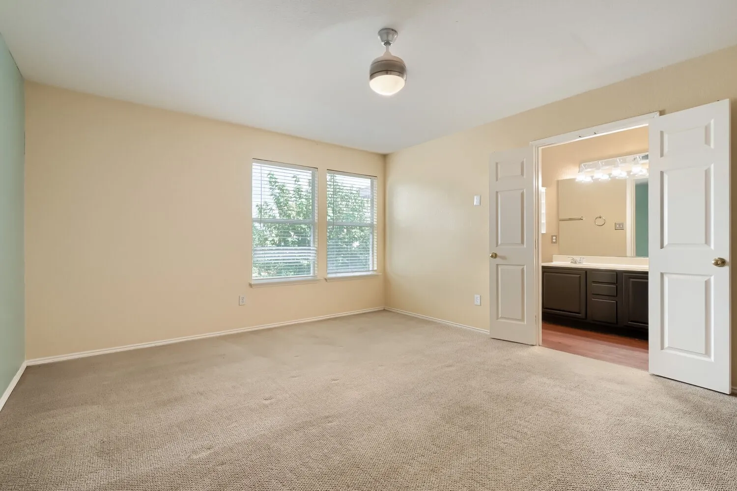 Spacious bedroom featuring neutral carpeting, two windows with blinds, a ceiling fan, and direct access to an attached bathroom