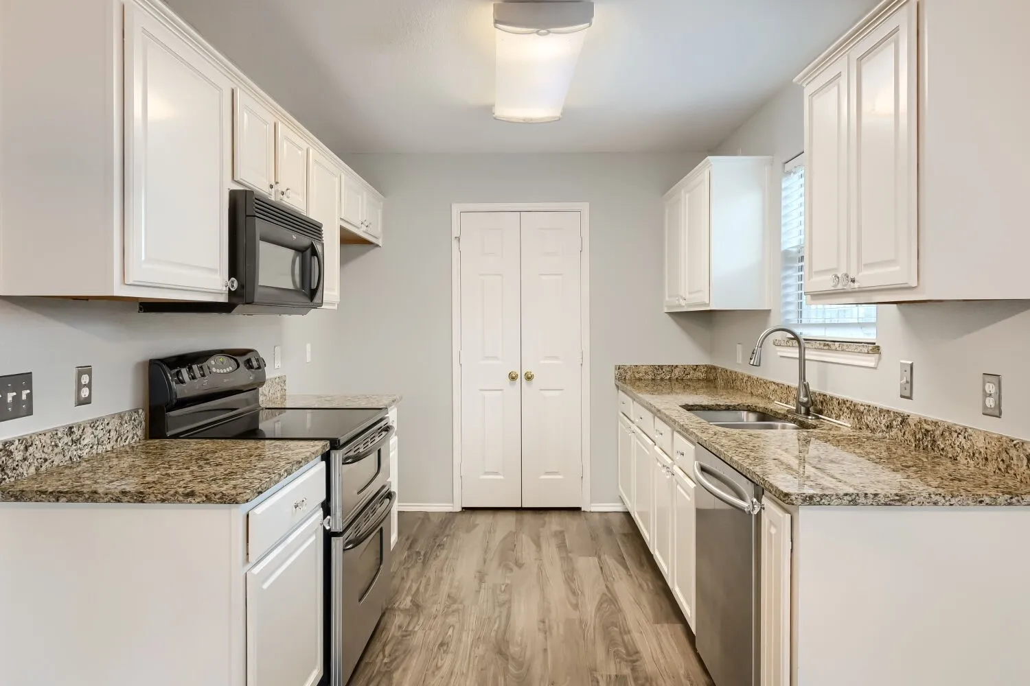 Galley kitchen featuring wood-finish flooring, white cabinetry, granite-style countertops, stainless steel sink, and a full suite of appliances