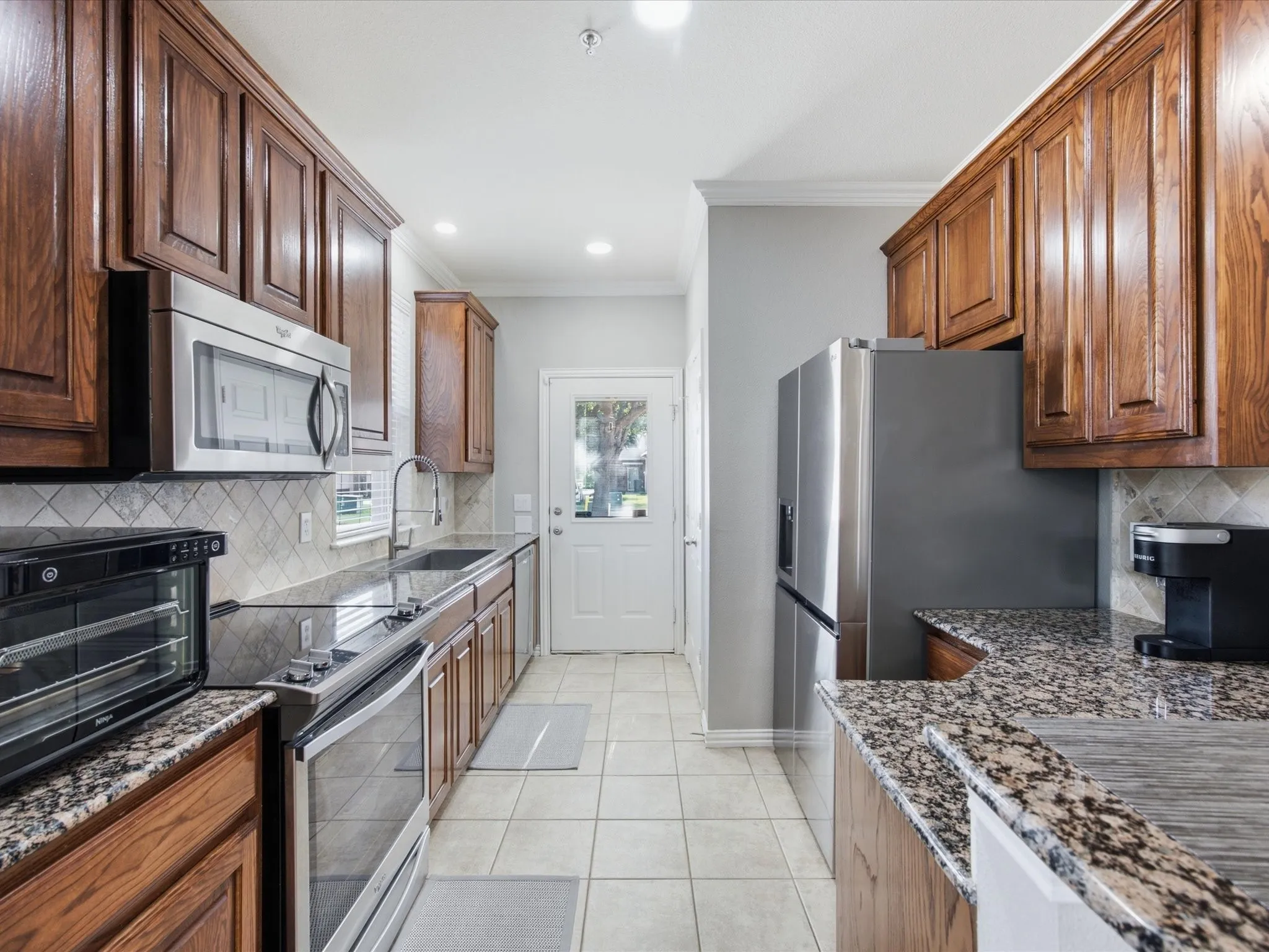 Galley kitchen featuring wood cabinetry, granite countertops, stainless steel appliances, a tile backsplash, and recessed lighting