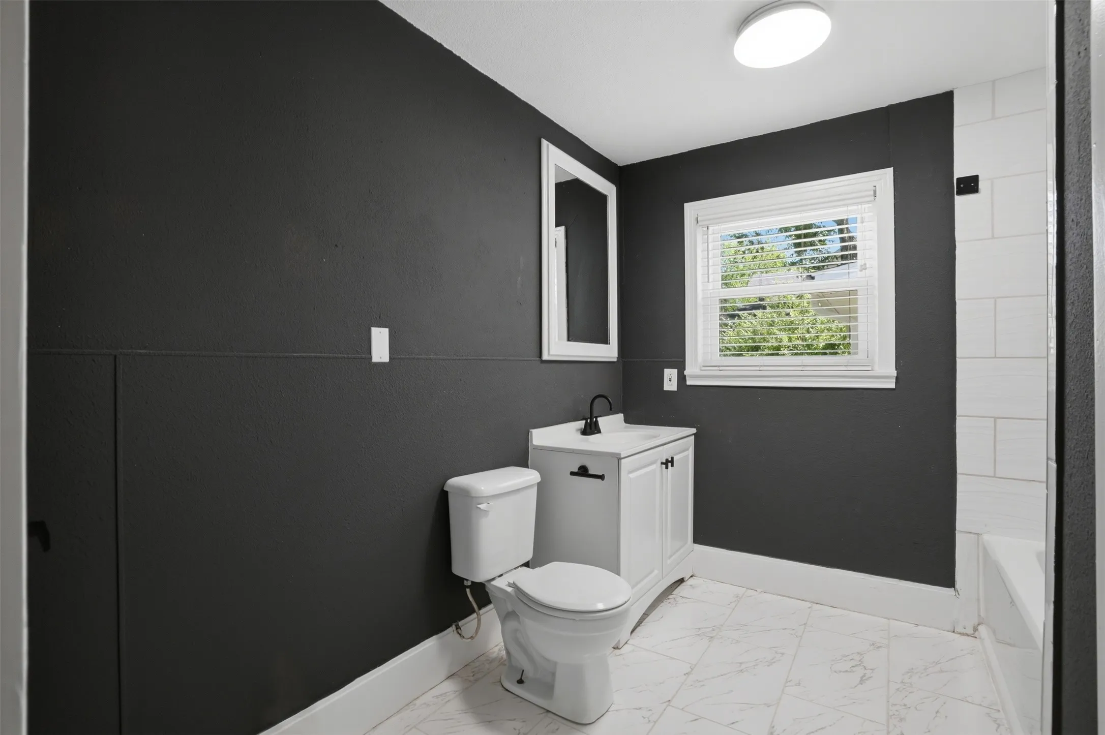 Bathroom featuring dark accent wall, white vanity with matte black hardware, white subway tile shower surround, and a window with blinds