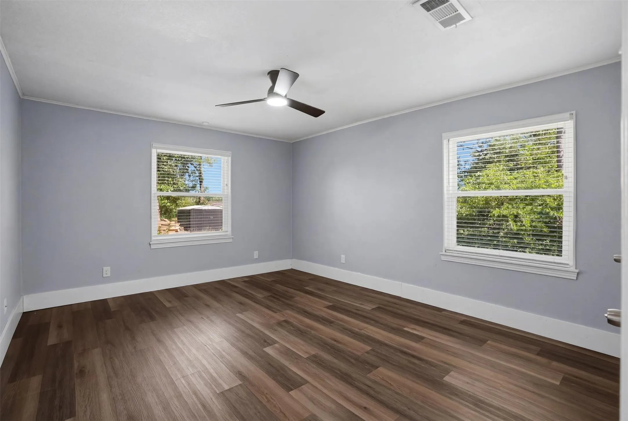Spacious room featuring wood-finish flooring, light blue walls, two large windows with blinds, a modern ceiling fan, and white crown molding
