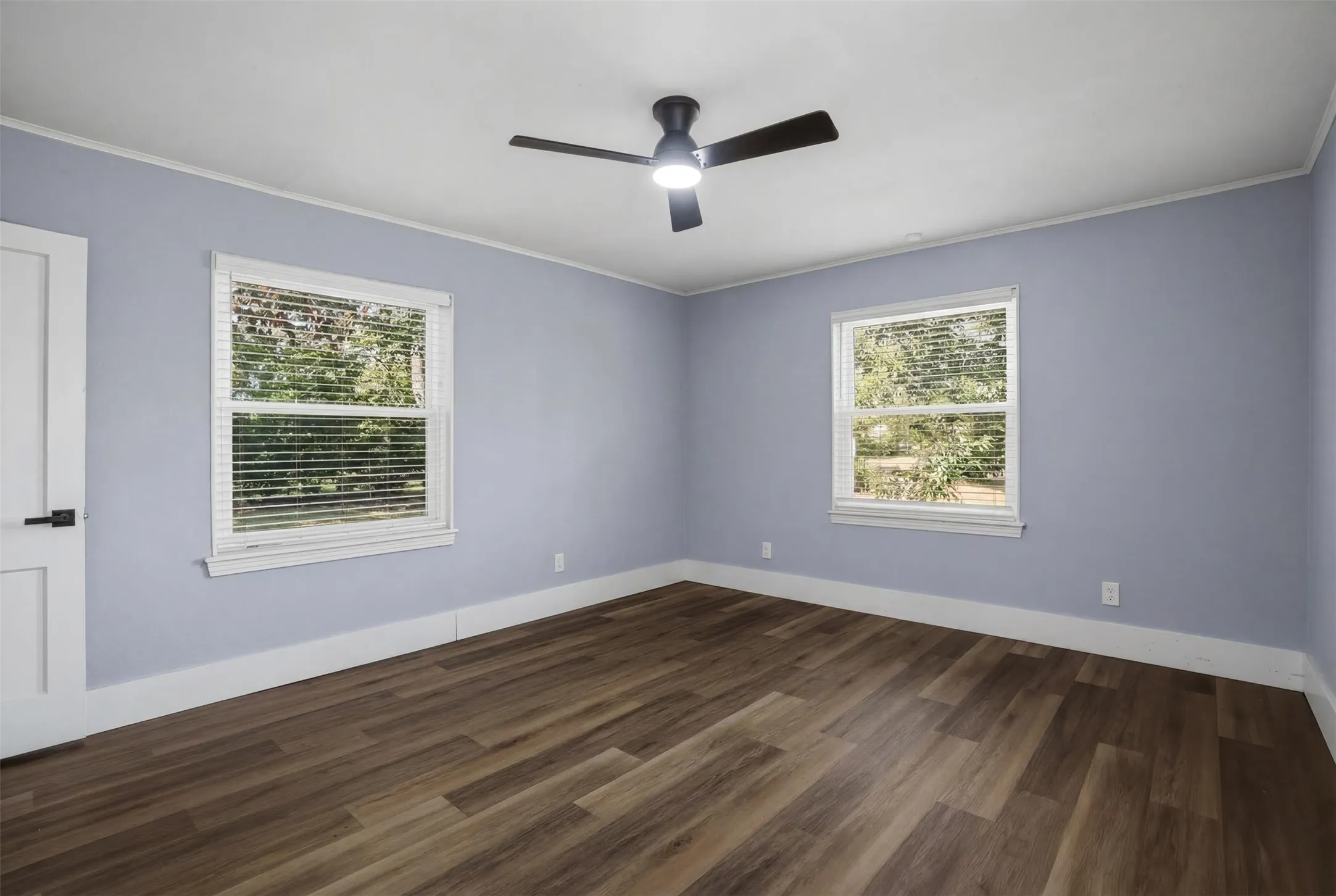 Spacious room featuring wood-finish flooring, two large windows with blinds, a modern ceiling fan with lighting, and a crisp white door with black hardware