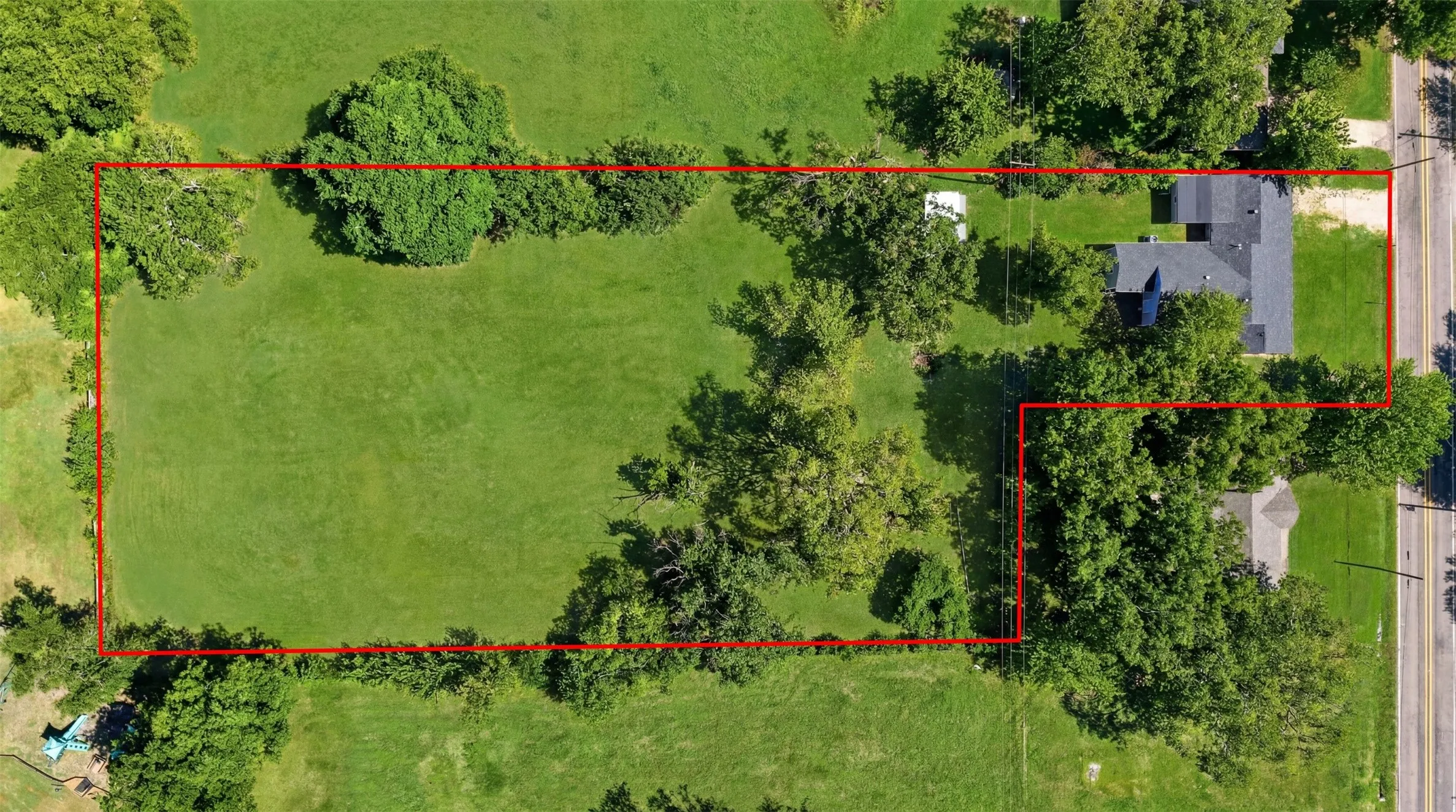 Expansive property showcasing a large grassy lot with mature trees, a single-story residence featuring a dark shingle roof, and direct access to a paved road