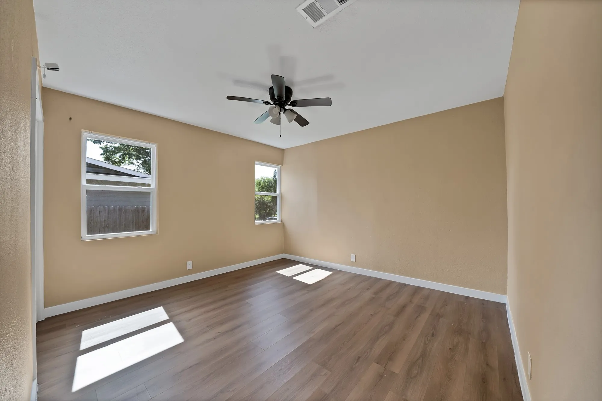 Spacious room featuring wood-finish flooring, two windows with white frames, a ceiling fan with integrated lighting, and neutral-toned walls