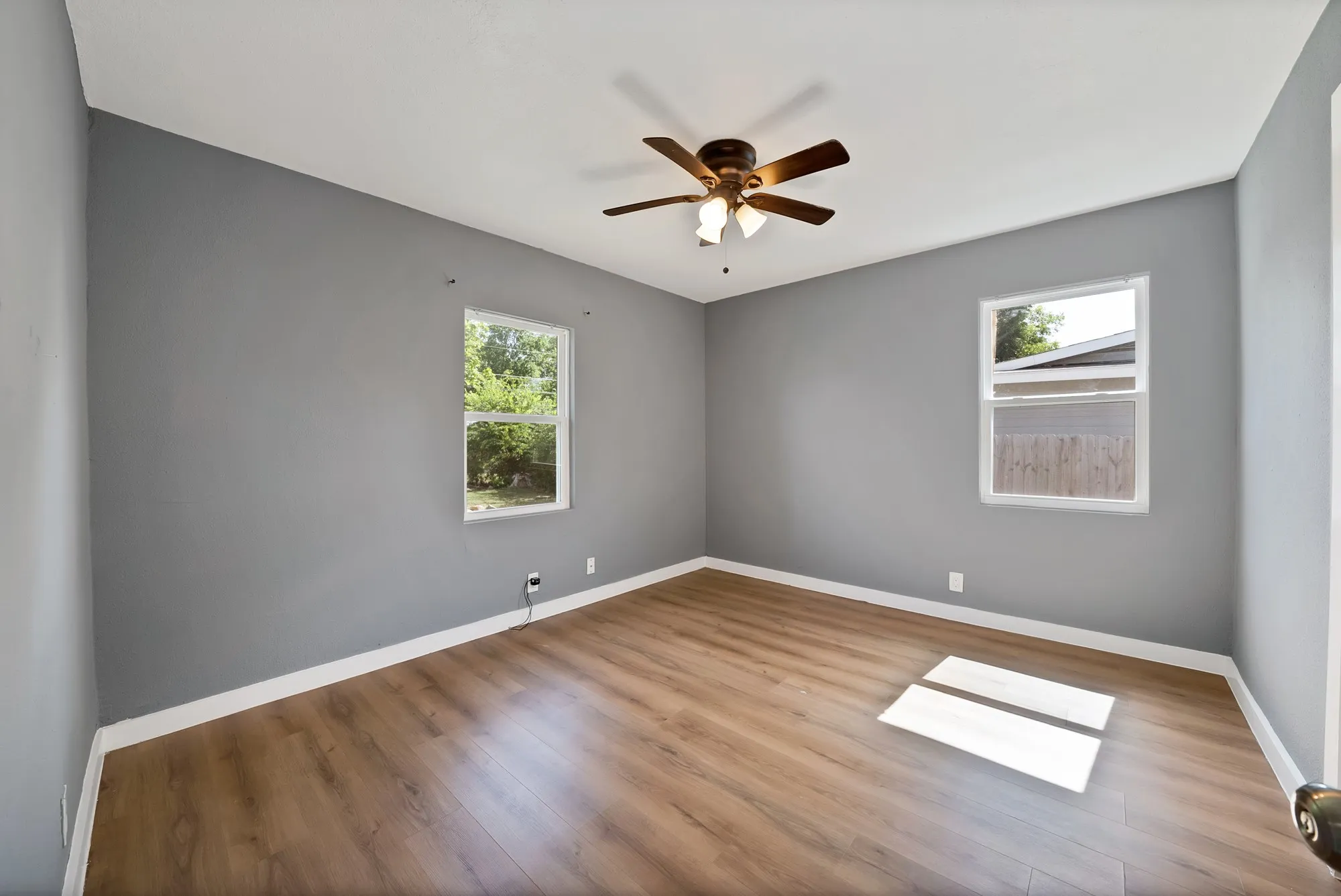 Spacious interior featuring wood-finish flooring, two windows, a ceiling fan with light fixture, and neutral gray walls