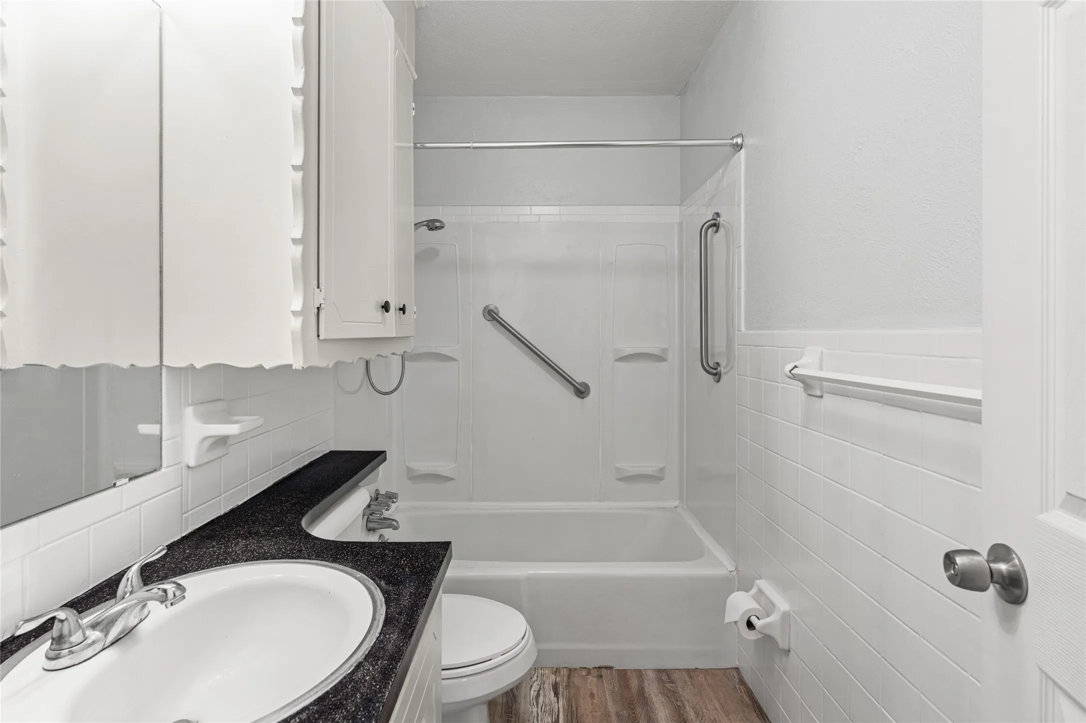 Full bathroom featuring a white tub/shower combination with grab bars, a vanity with a dark countertop and undermount sink, a toilet, and wood-finish flooring