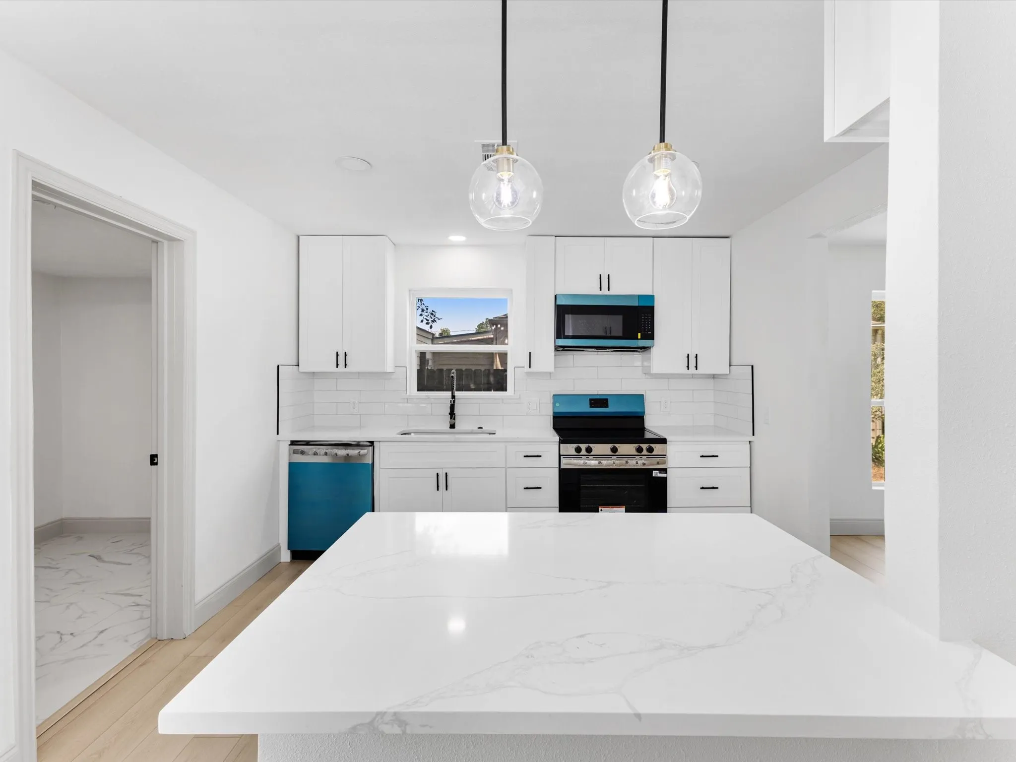 Bright kitchen featuring a substantial island with a white veined countertop, white shaker cabinetry, stainless steel appliances with blue accents, a white subway tile backsplash, and light wood-finish flooring