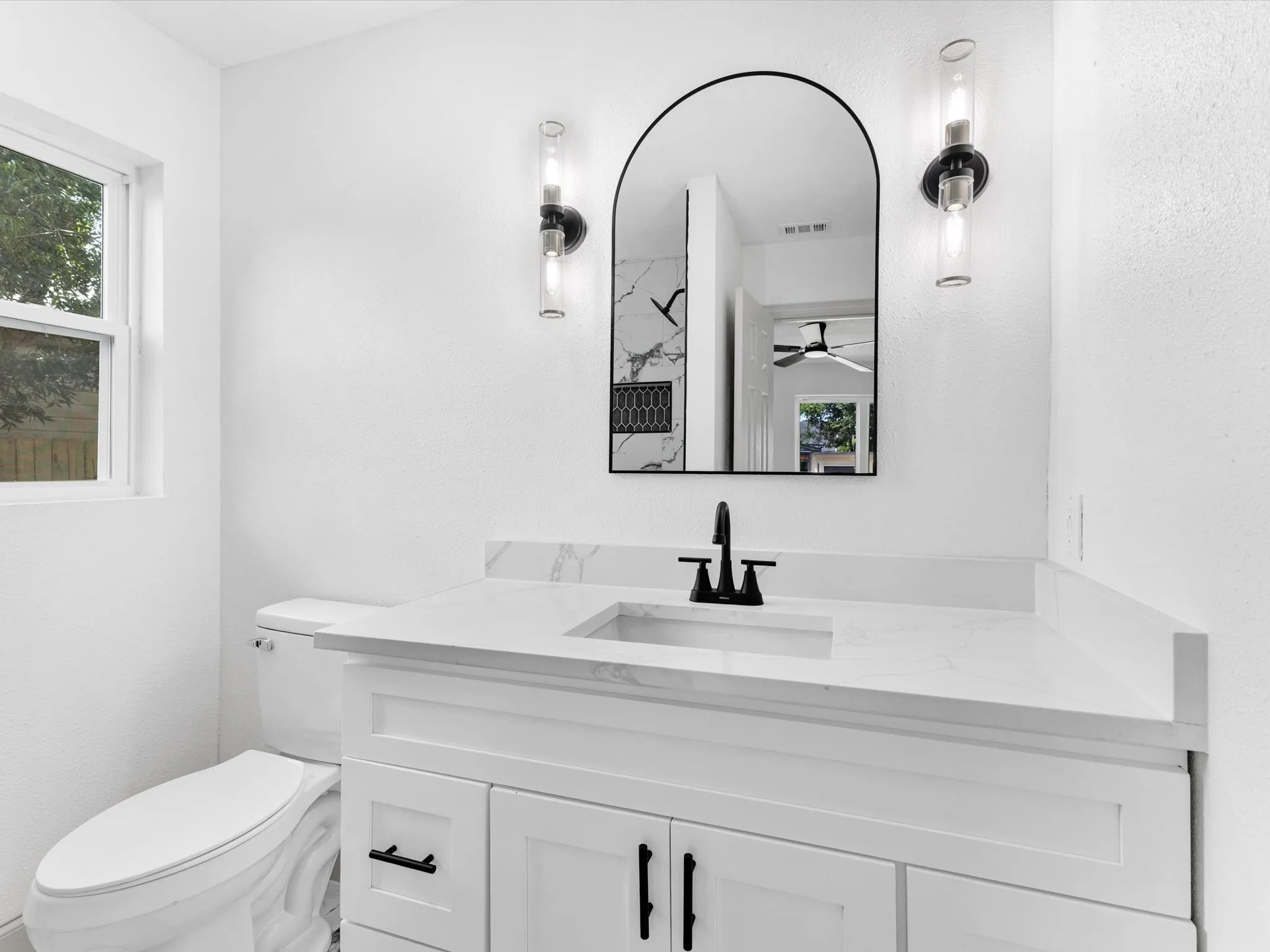Bathroom featuring a white vanity with panel cabinetry, a white countertop, a black faucet, and an arched mirror with sconce lighting