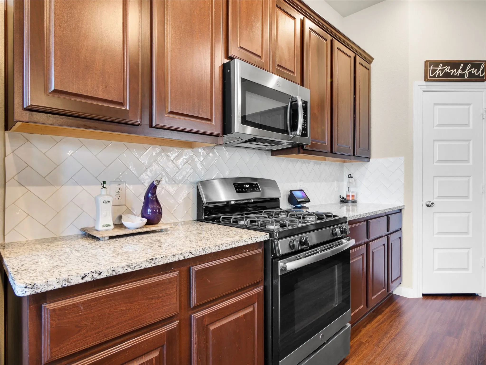 Kitchen featuring wood-finish cabinetry, stainless steel microwave, gas range, granite-style countertops, and a white herringbone tile backsplash
