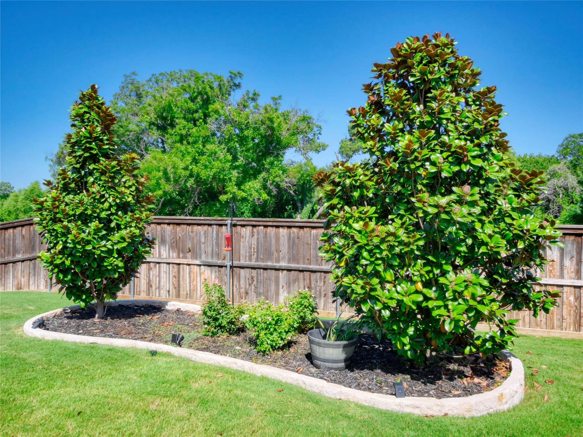 Expansive grassy yard featuring a wood privacy fence, established trees, and curved stone-edged garden beds with dark mulch
