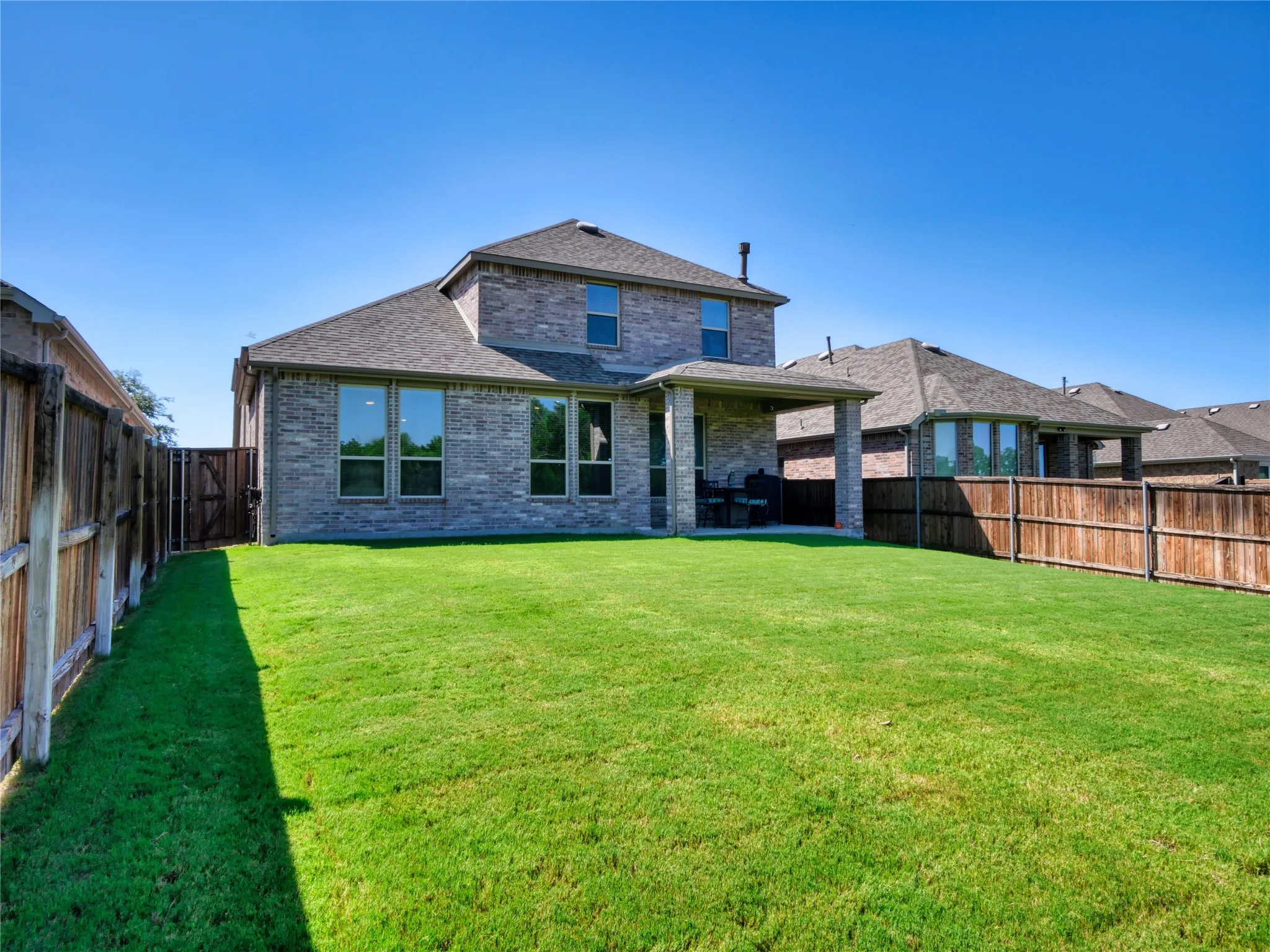 Expansive green lawn with a covered patio, brick exterior, multi-pane windows, and a wood fence