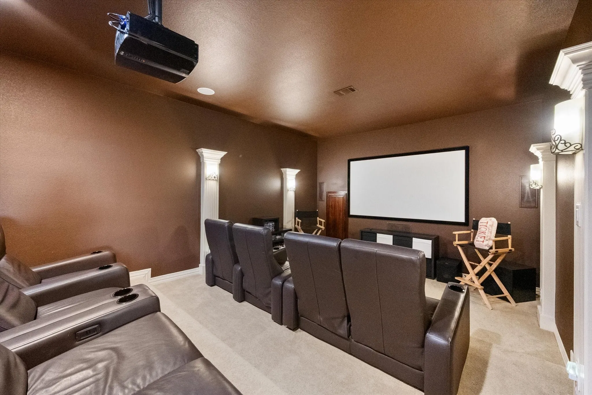 Dedicated media room featuring a large projection screen, ceiling-mounted projector, wall sconces, decorative architectural columns, and carpet flooring