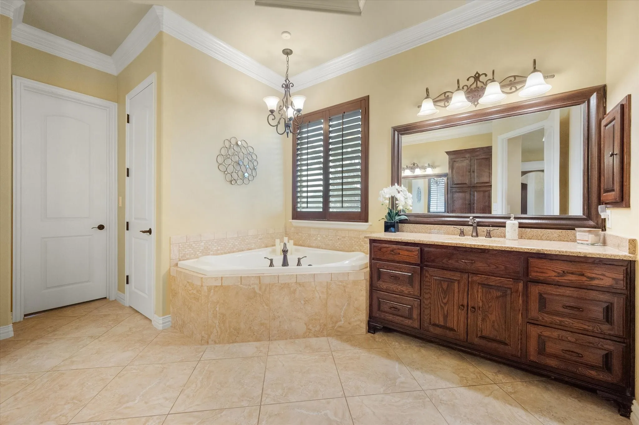 Spacious bathroom featuring a corner soaking tub with tile surround, a large dark wood vanity with ample storage, a light-colored countertop, and a wide framed mirror with decorative lighting