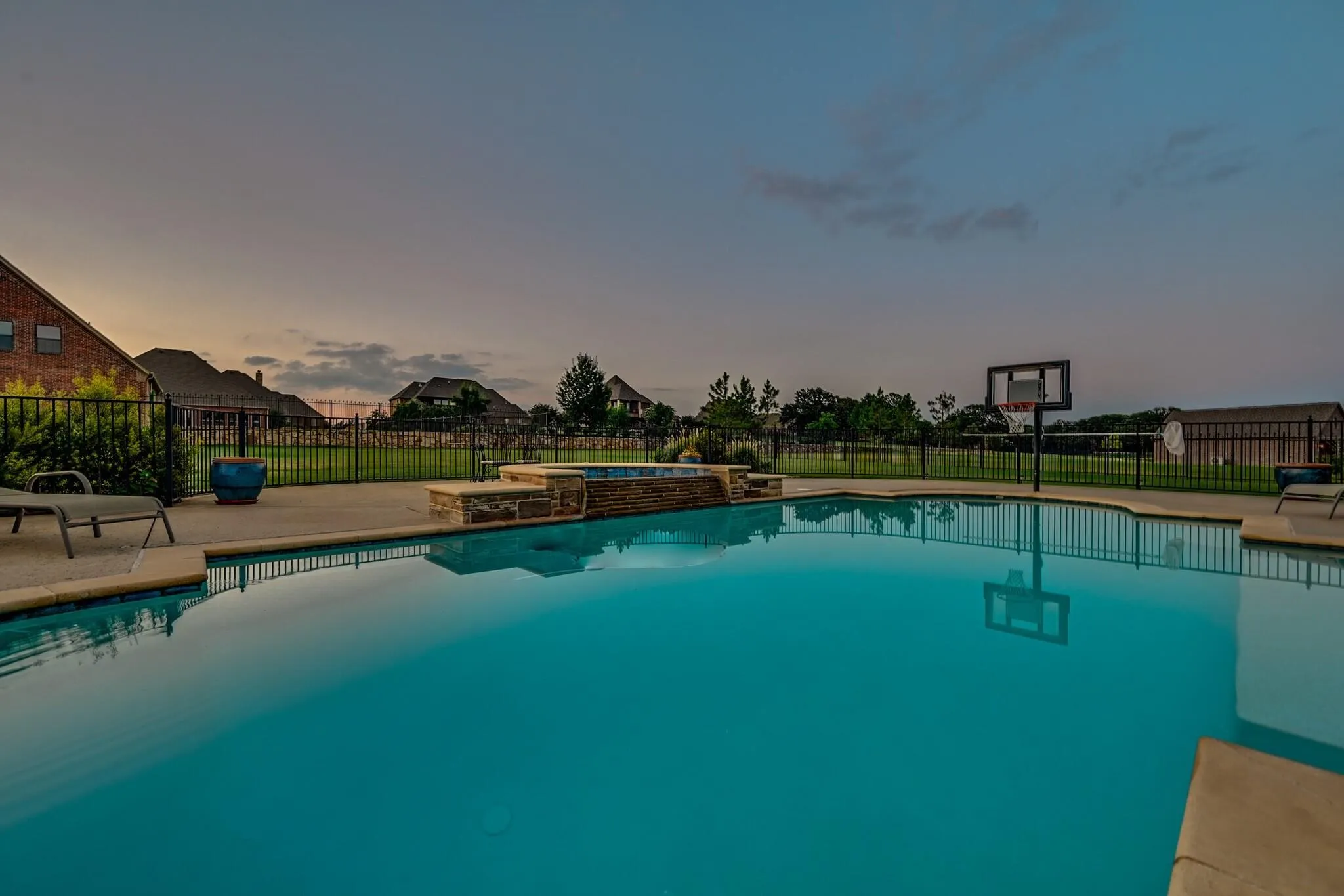 In-ground swimming pool featuring a built-in spa with stone surround, concrete decking, and a perimeter fence