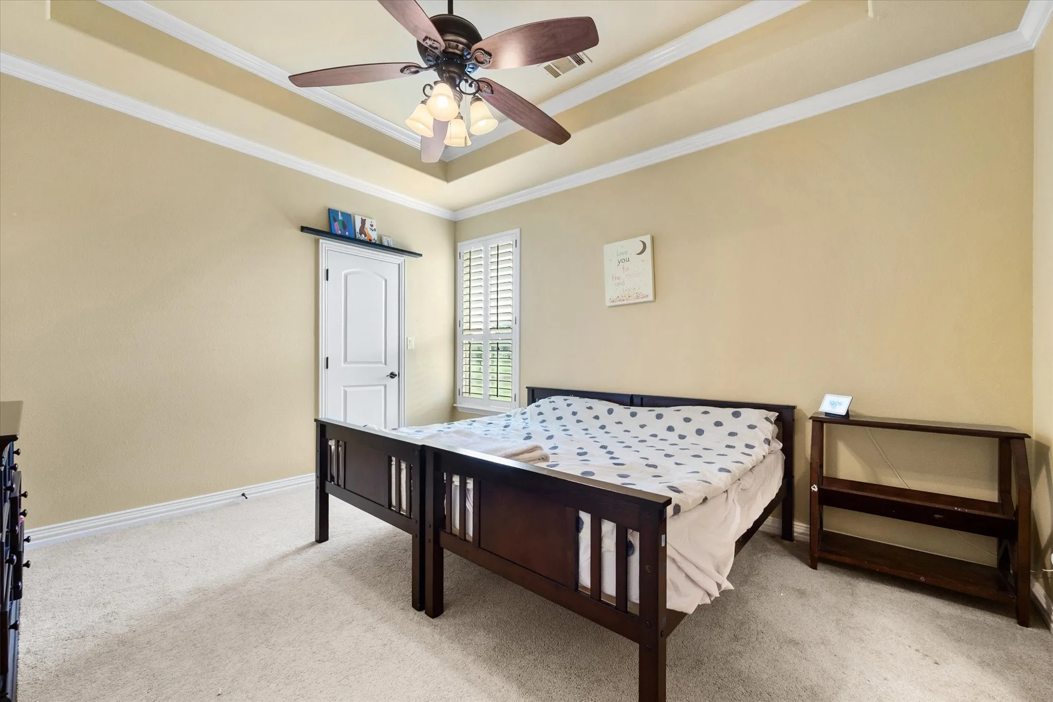 Recessed ceiling with crown molding, ceiling fan with light fixture, carpeted flooring, window with plantation shutters, and a paneled door
