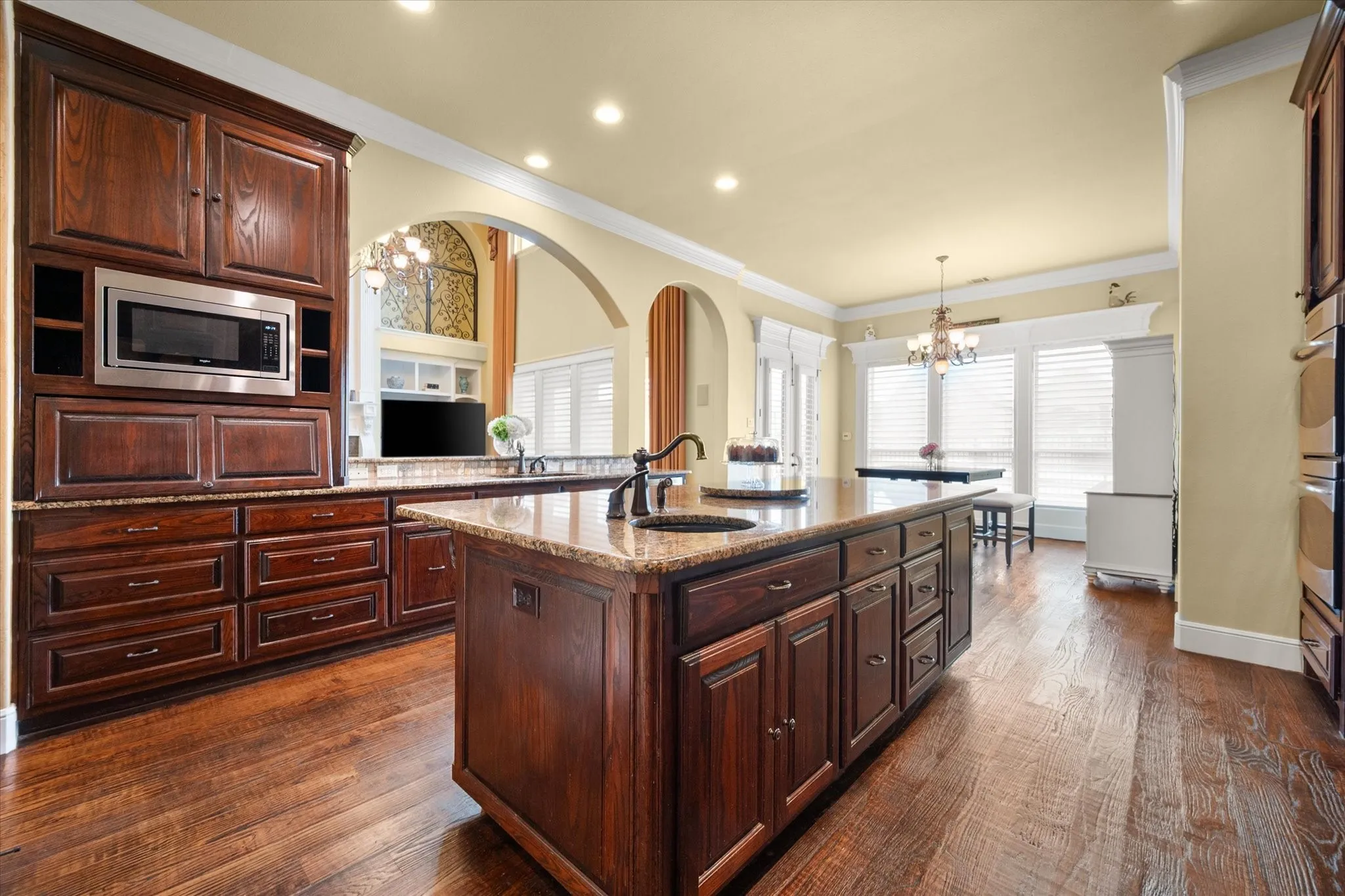 Spacious kitchen featuring a large central island with an undermount sink, rich wood-finish cabinetry, stone countertops, integrated stainless steel appliances, and wood-finish flooring