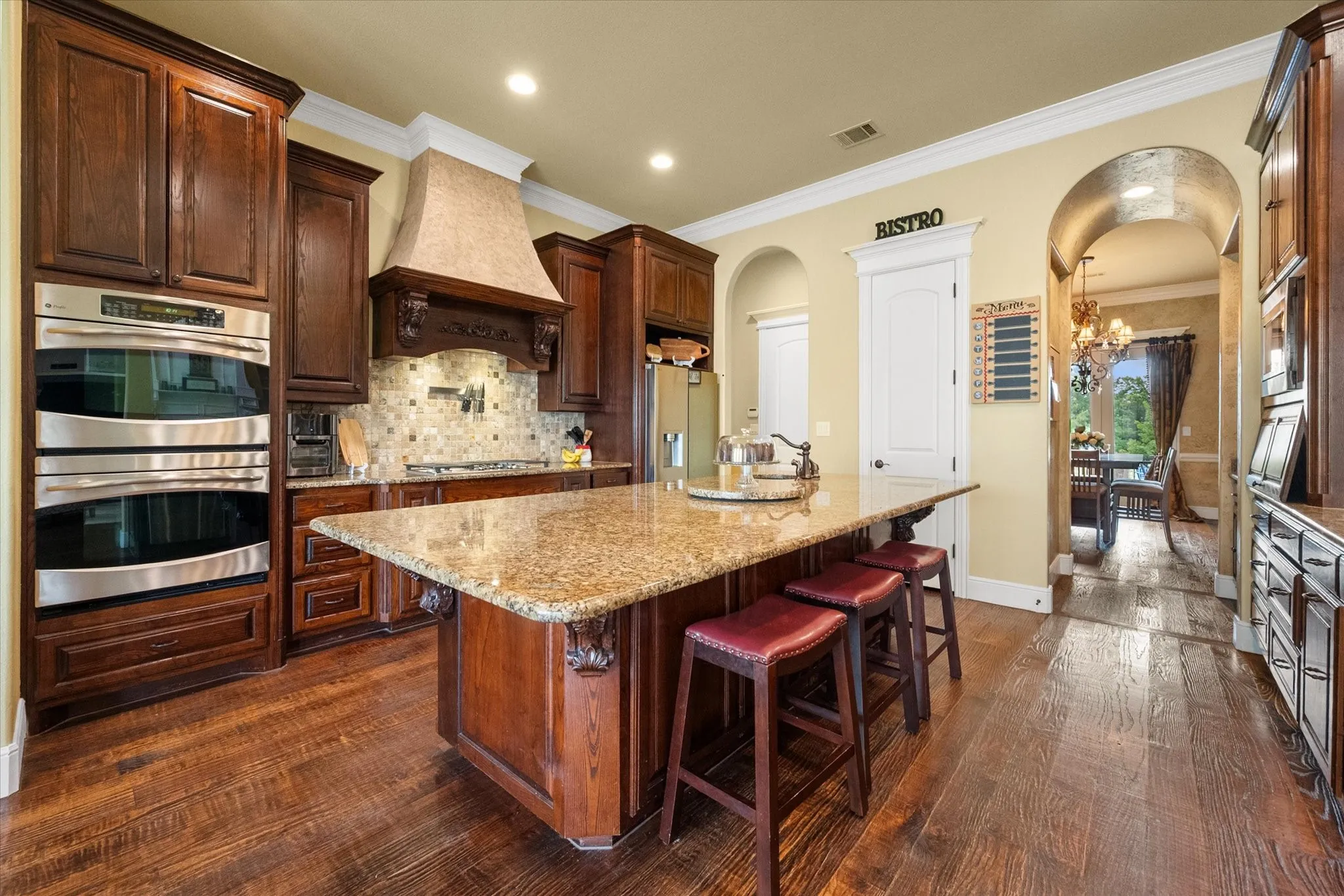 Gourmet kitchen with wood-finish flooring, dark wood cabinetry, stainless steel double ovens, a stone-finish backsplash, and a large central island with a granite countertop