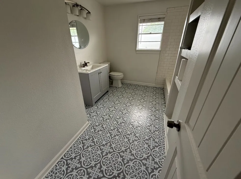 Bathroom featuring patterned floor tiles, a single window, subway tile shower surround, a gray vanity with an integrated sink, and a circular wall mirror