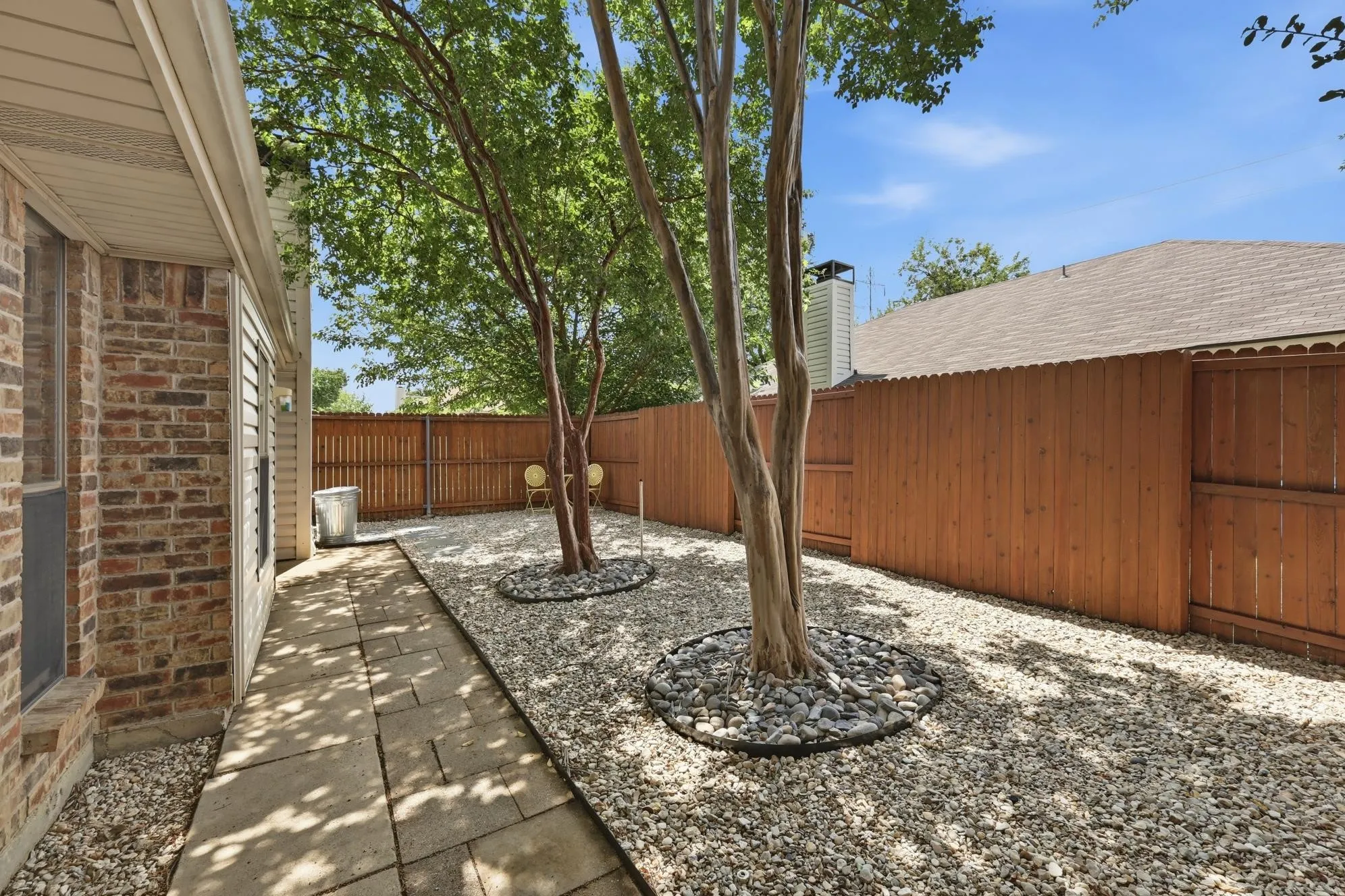Private side yard featuring a stone paver walkway, mature trees within rock beds, and a wood privacy fence