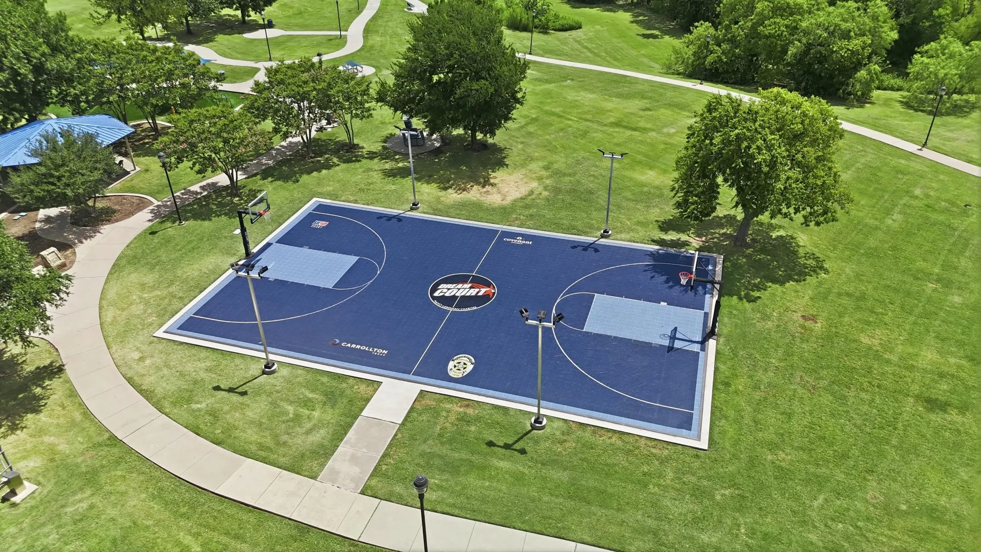 Full-size outdoor basketball court featuring a blue playing surface with light blue key areas, complete with hoops and integrated lighting.