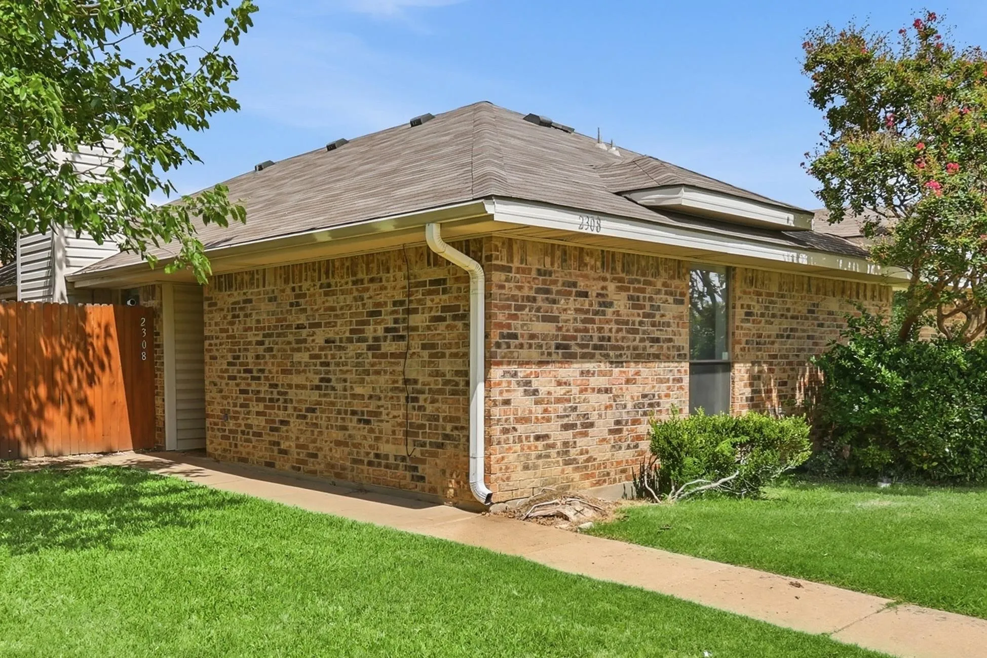 Brick exterior with a brown shingle roof, featuring a concrete pathway, mature green lawn, and a wooden privacy fence