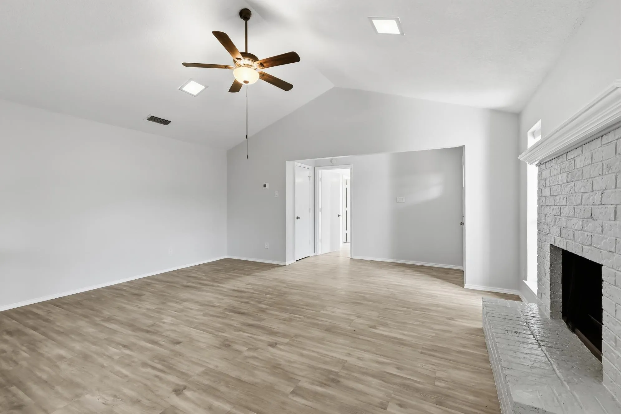 Spacious living area featuring a vaulted ceiling with a ceiling fan, recessed lighting, wood-finish flooring, a painted brick fireplace with hearth, and an open doorway