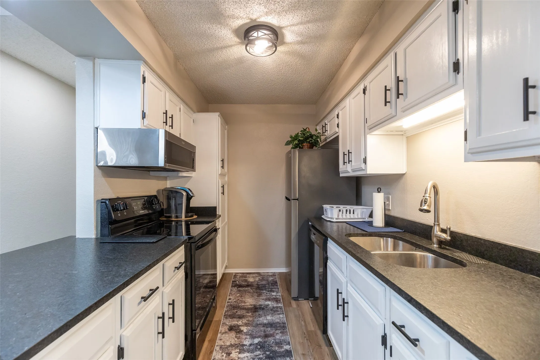 Galley kitchen featuring white cabinetry, dark countertops, black appliances, stainless steel sink, and wood-finish flooring