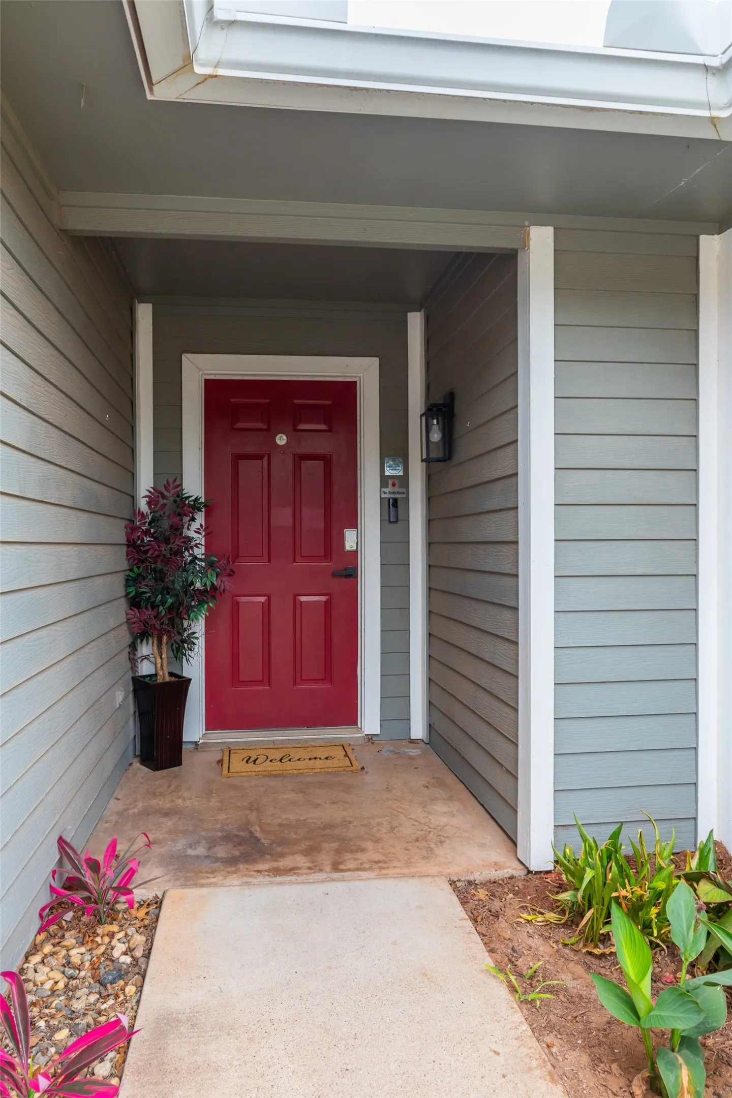 Entryway featuring a bold red paneled door, light gray horizontal siding, an exterior sconce light, and a concrete path
