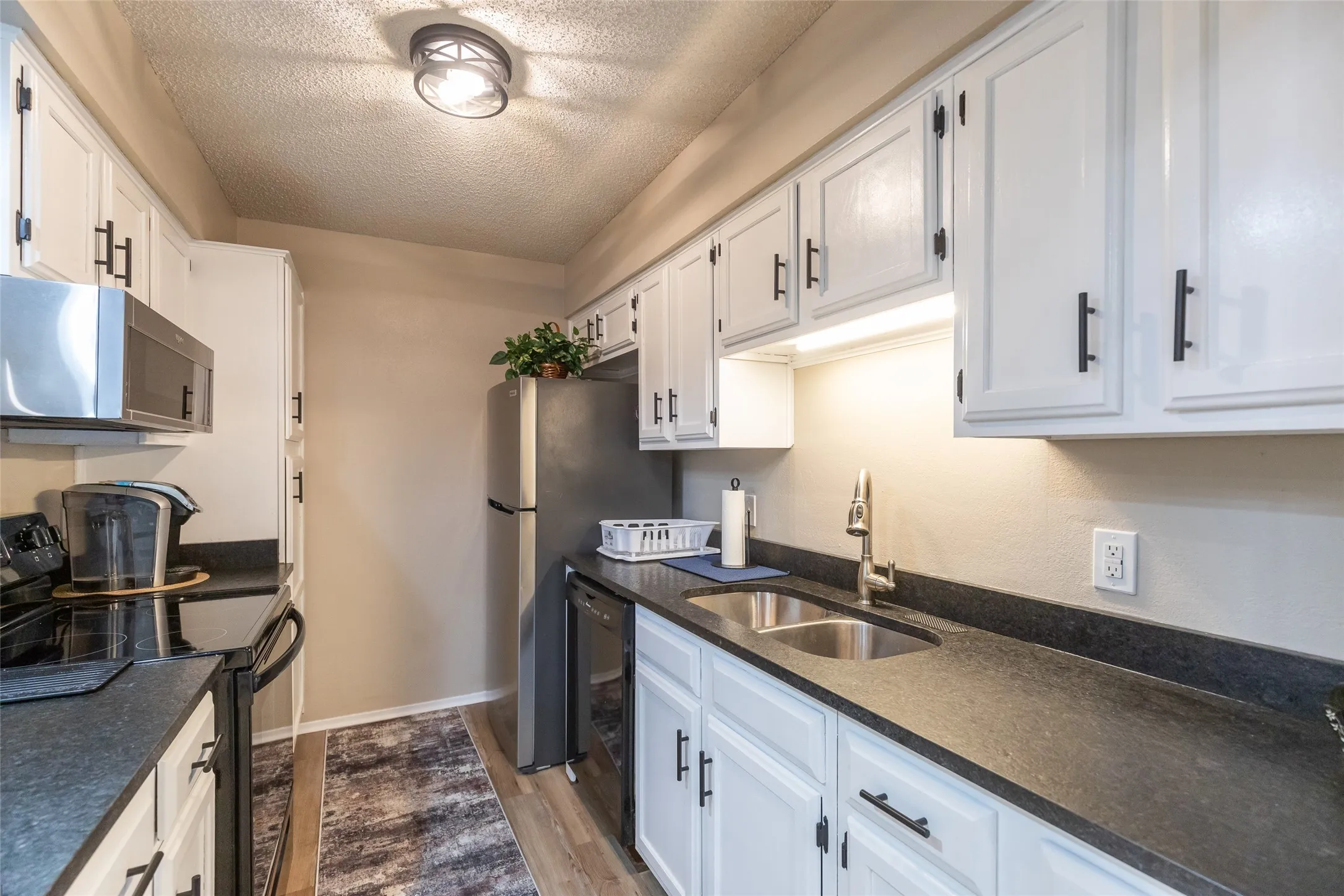 Galley kitchen featuring white cabinetry with matte black hardware, dark countertops, under-cabinet lighting, wood-finish flooring, and stainless steel appliances including a built-in microwave