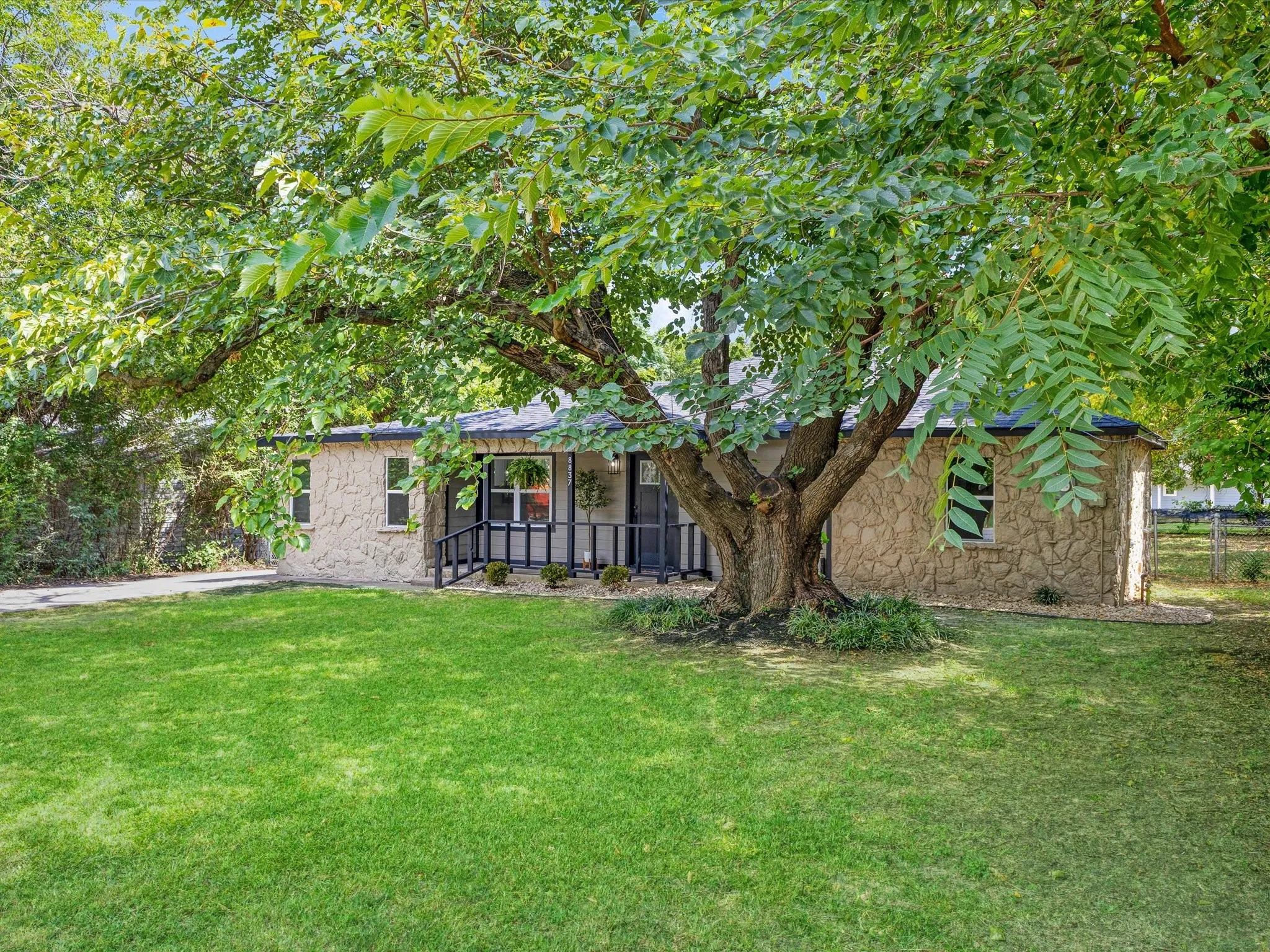 Front exterior featuring natural stone siding, a covered porch with black railing, a mature shade tree, a manicured lawn, and a paved driveway
