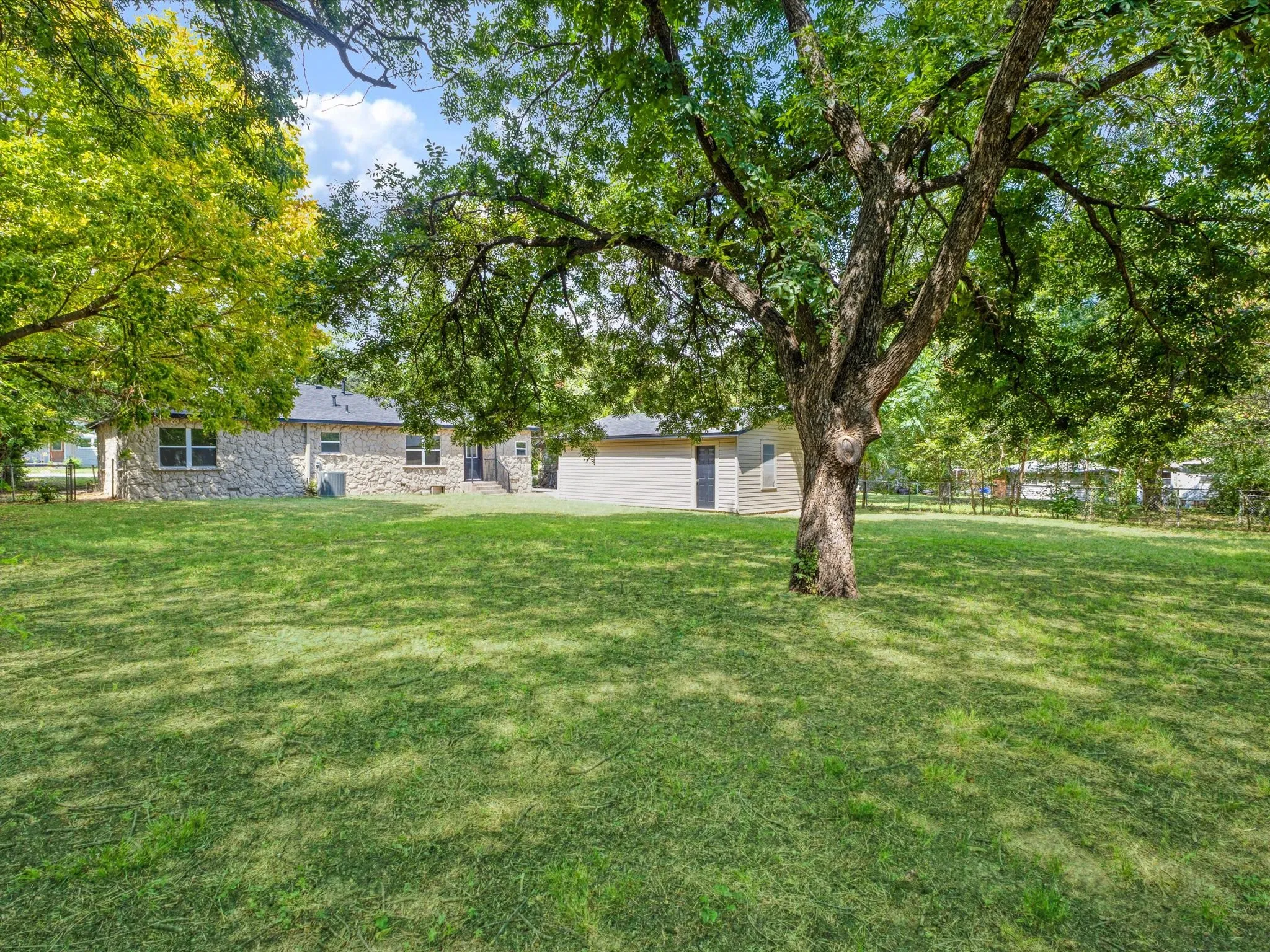Spacious backyard featuring a substantial mature tree, expansive lawn, and a stone-clad exterior on the main residence