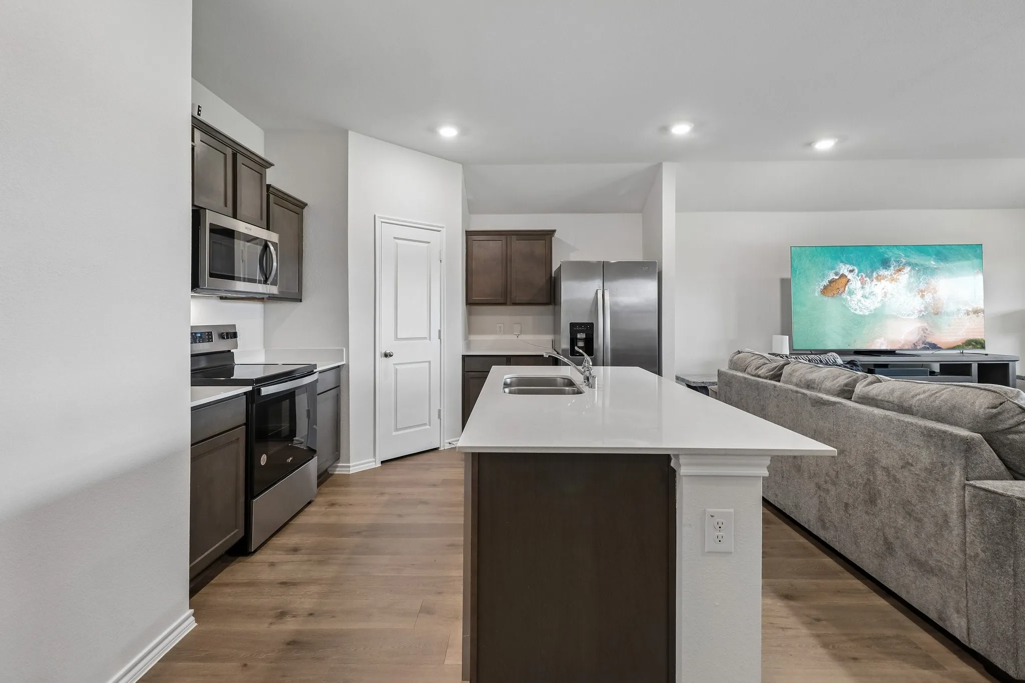 Modern kitchen featuring wood-finish flooring, dark wood cabinetry, stainless steel appliances, a kitchen island with sink, and light countertops