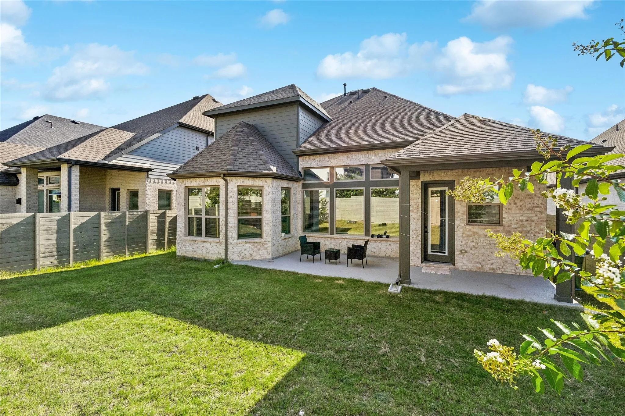 Expansive backyard with a manicured lawn, featuring a covered patio, brick exterior, bay windows, and a privacy fence
