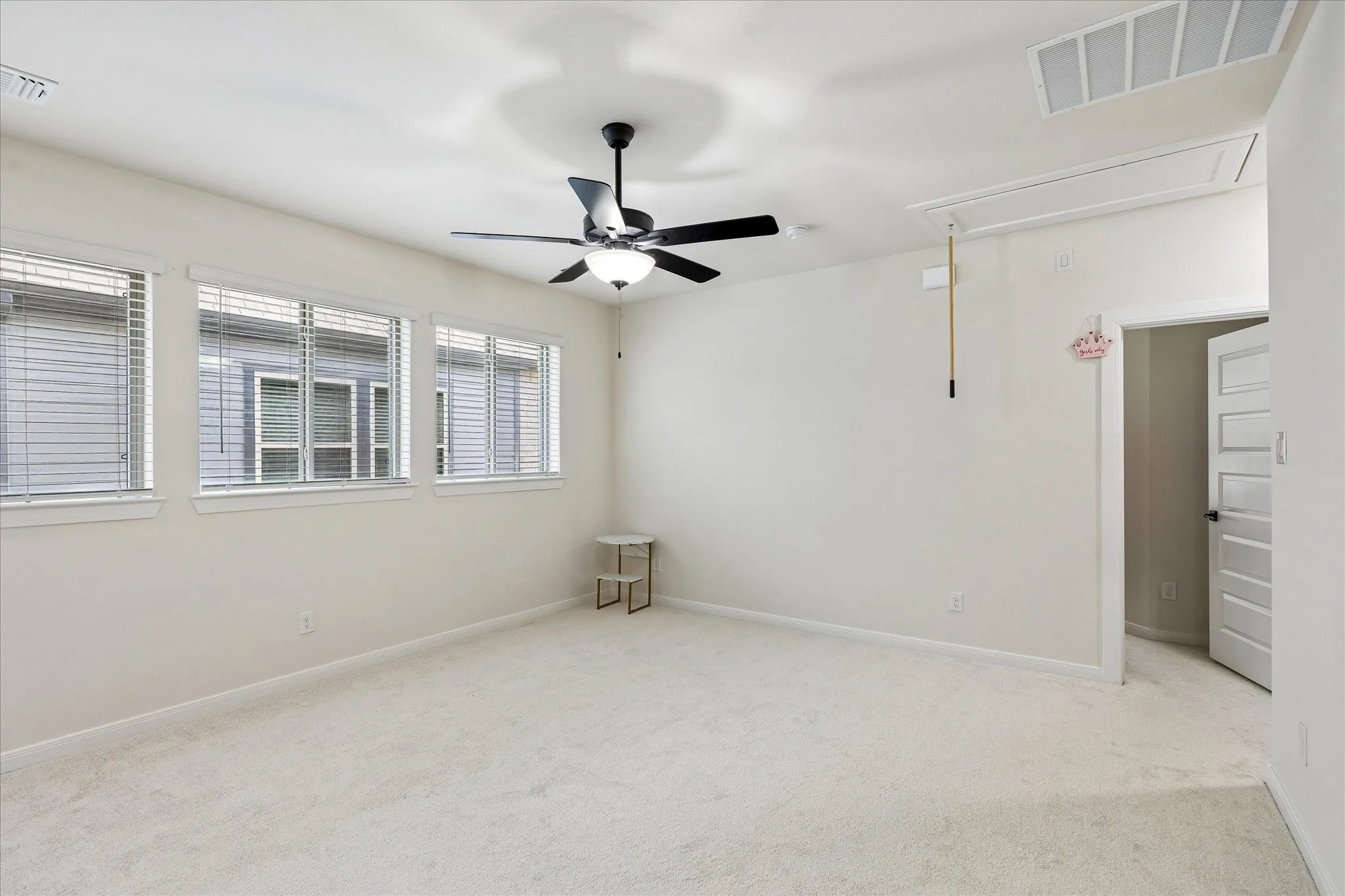 Expansive carpeted room featuring a ceiling fan with integrated lighting, three windows with blinds, a white paneled door, and an overhead attic access panel