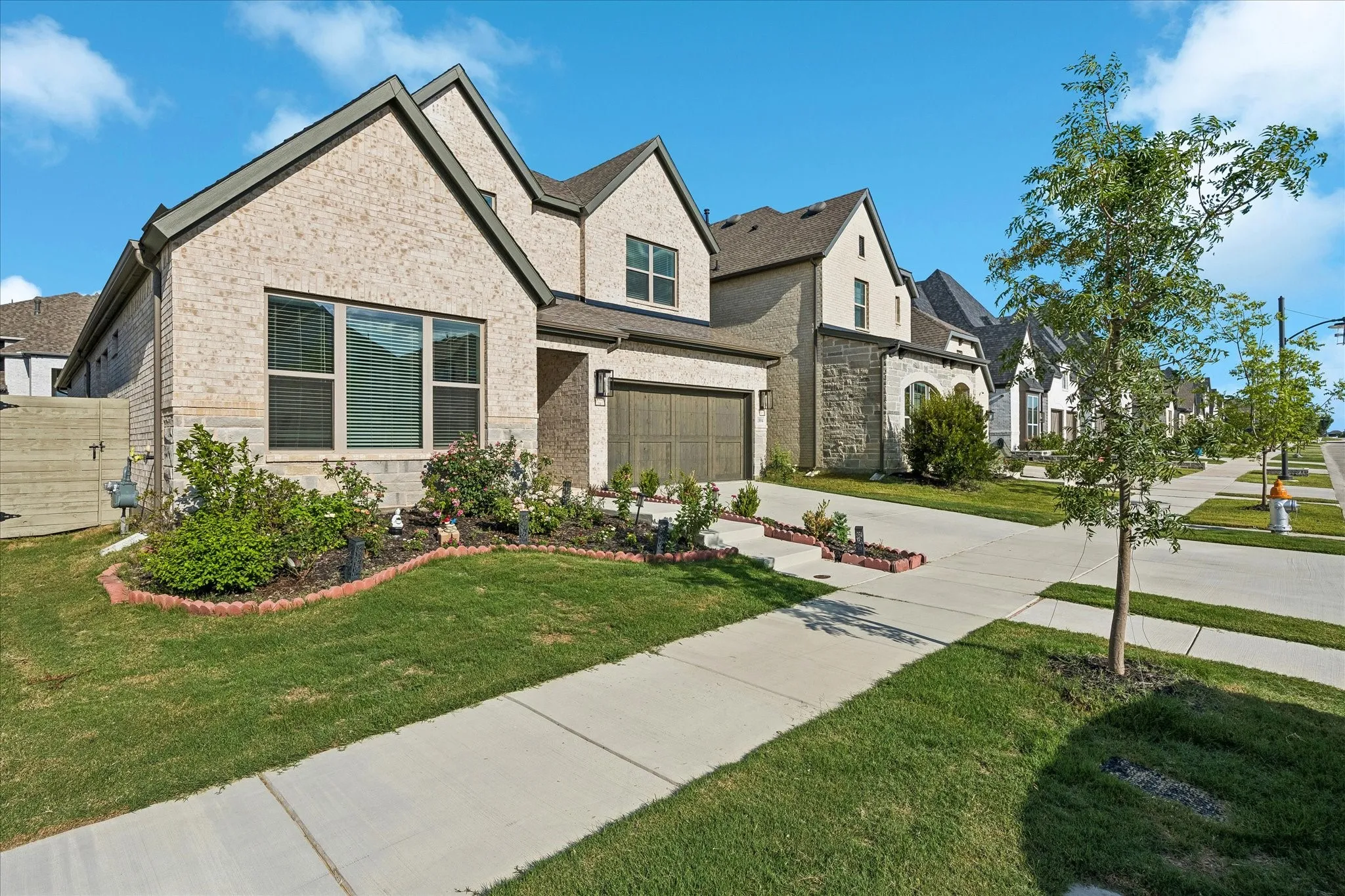 Contemporary exterior featuring light brick, dark trim, an attached garage with a wood-finish door, a manicured lawn, and mature landscaping