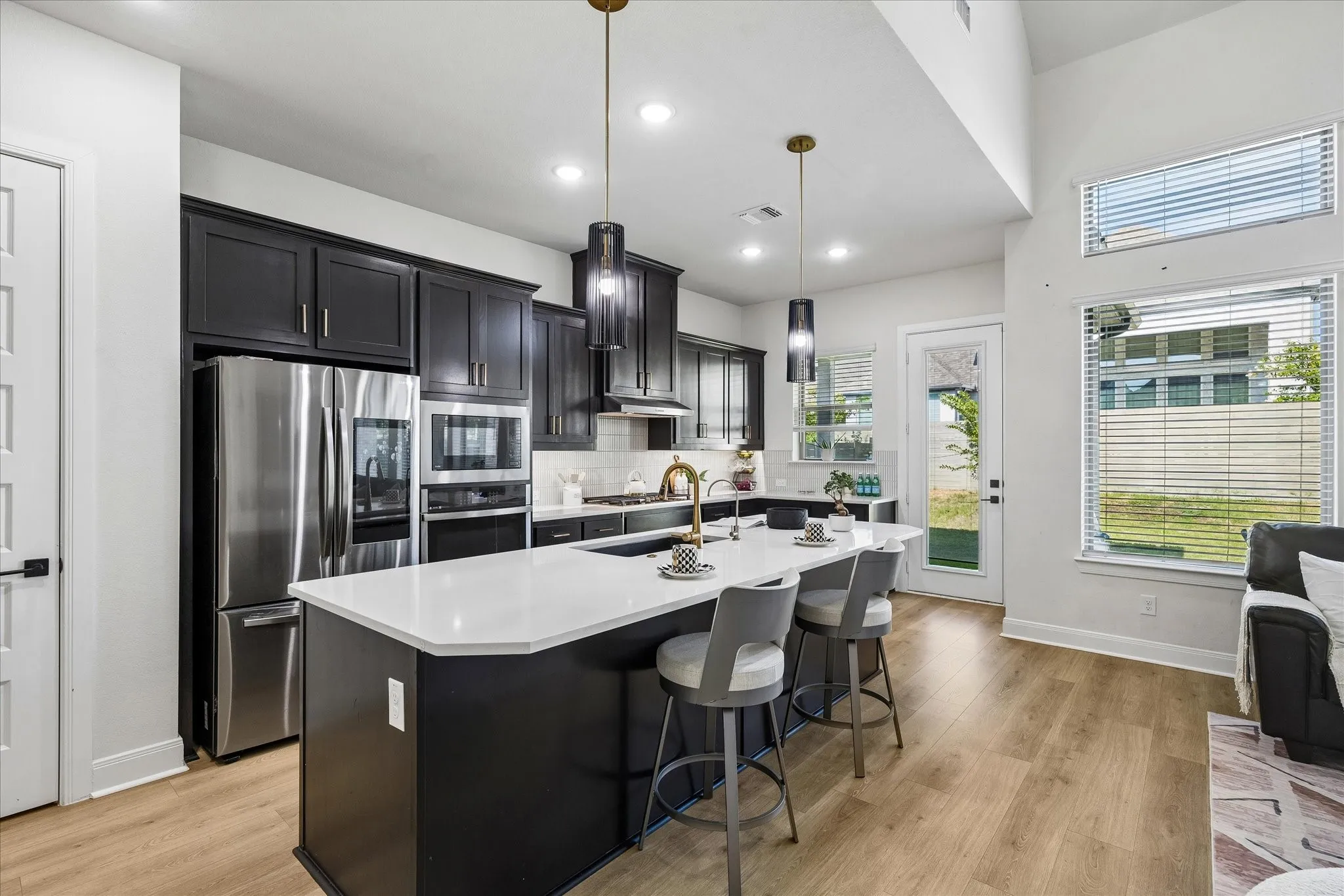 Open-concept kitchen featuring dark cabinetry, stainless steel appliances, a central island with a light-toned countertop, and gold-finish pendant lighting