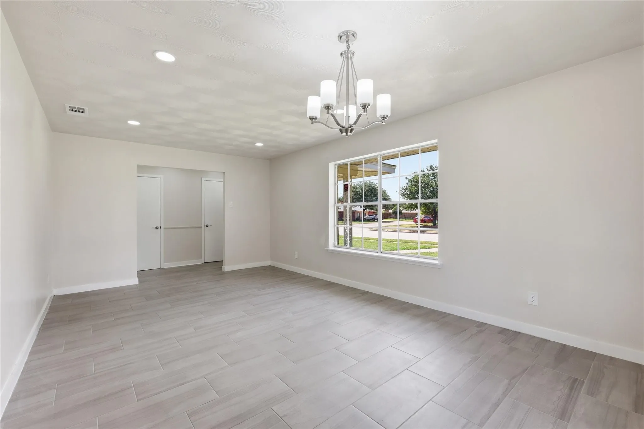 Spacious room featuring light gray walls, large front-facing window, recessed lighting, and a chrome chandelier with five frosted glass shades