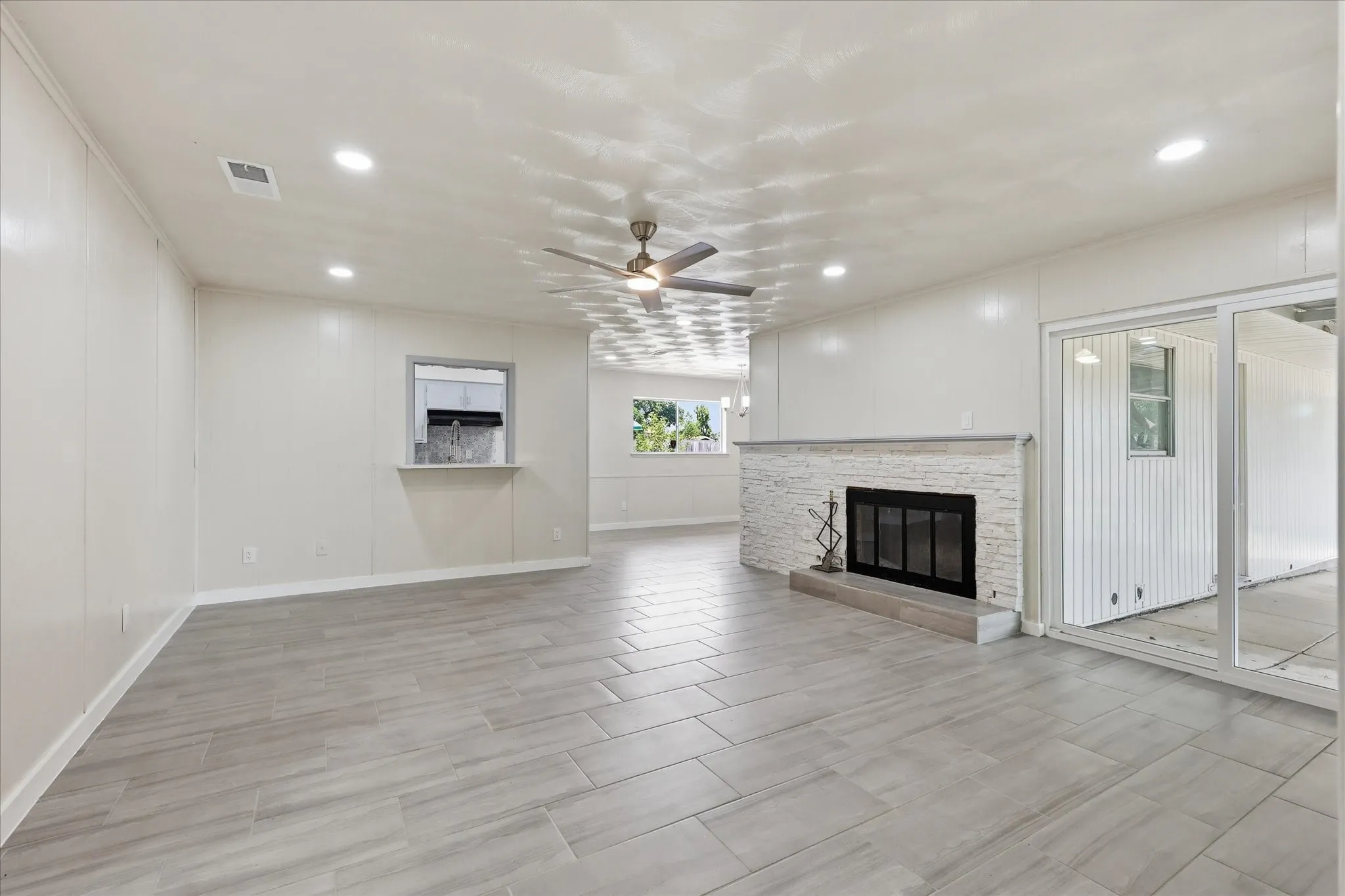 Spacious living area featuring light wood-finish flooring, a white brick fireplace with a dark firebox, a ceiling fan, recessed lighting, and a pass-through window with a lower shelf