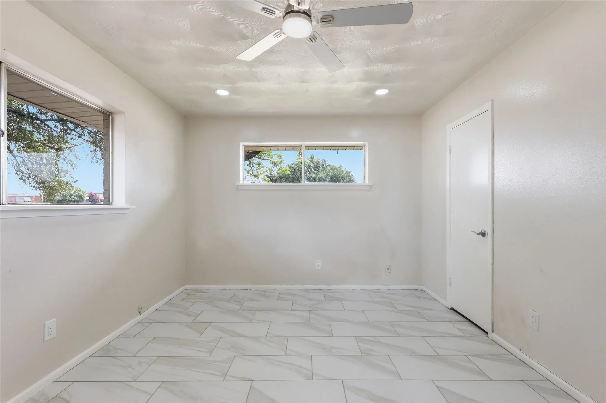 Spacious room featuring large format tile flooring, two windows, recessed lighting, a ceiling fan, and a solid door