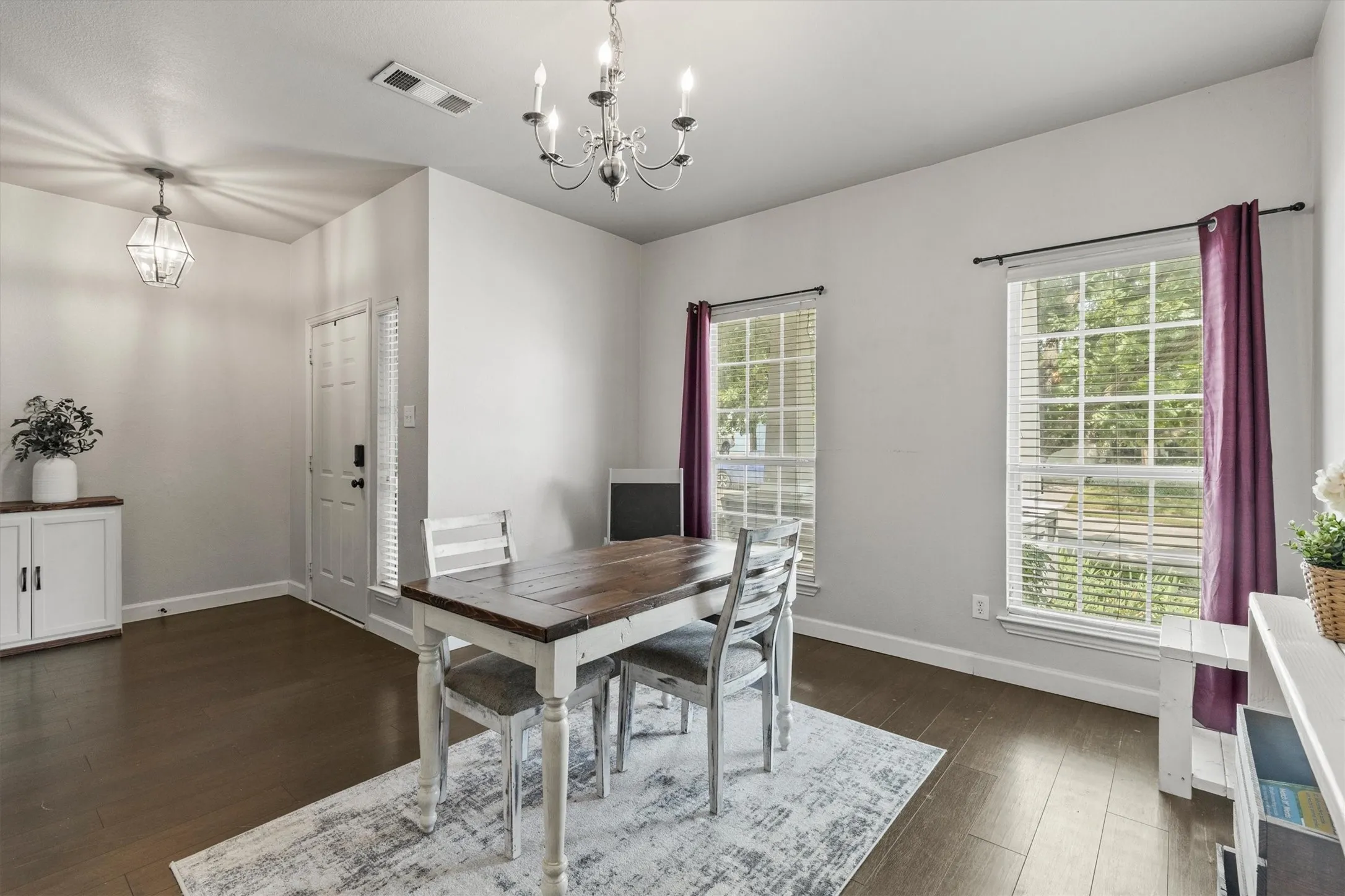 Open-concept dining space featuring wood-finish flooring, a contemporary chandelier, two large windows with blinds, and white baseboards