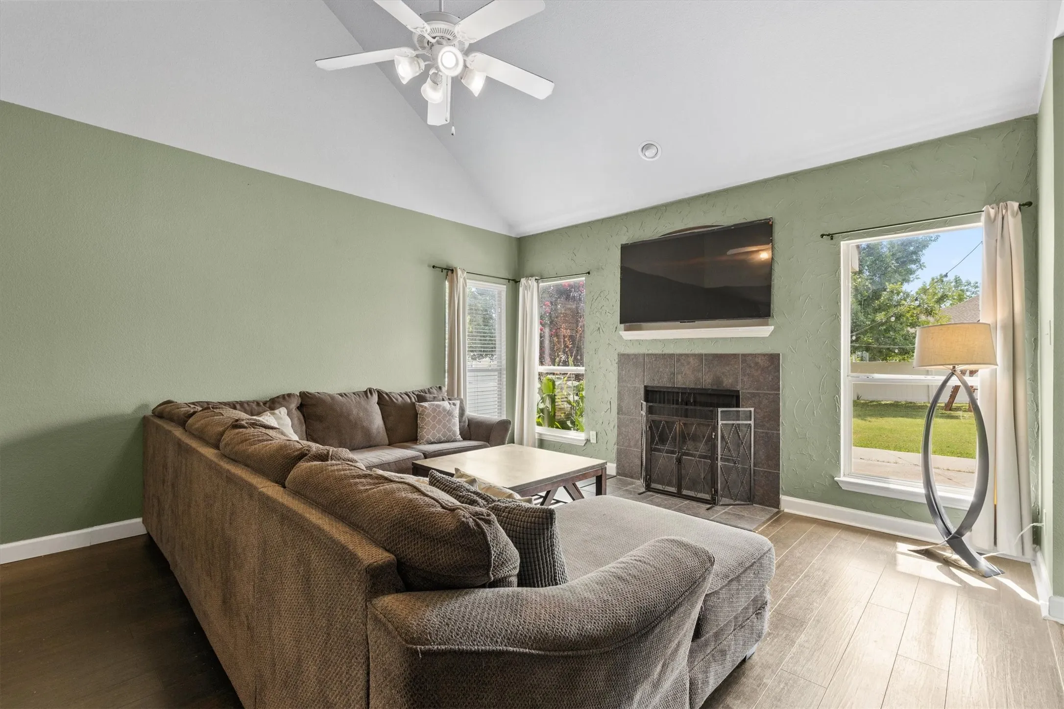 Vaulted ceiling living area featuring wood-finish flooring, a tiled fireplace with mantel, and multiple windows