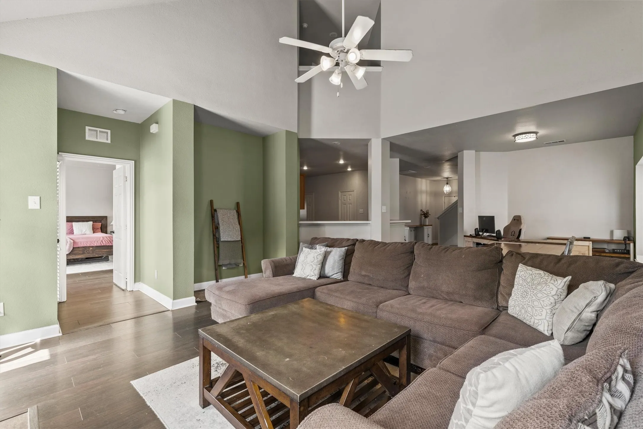 Spacious living area featuring high vaulted ceilings, a ceiling fan with integrated lighting, wood-finish flooring, a pass-through opening to another room, and a doorway leading to a separate space