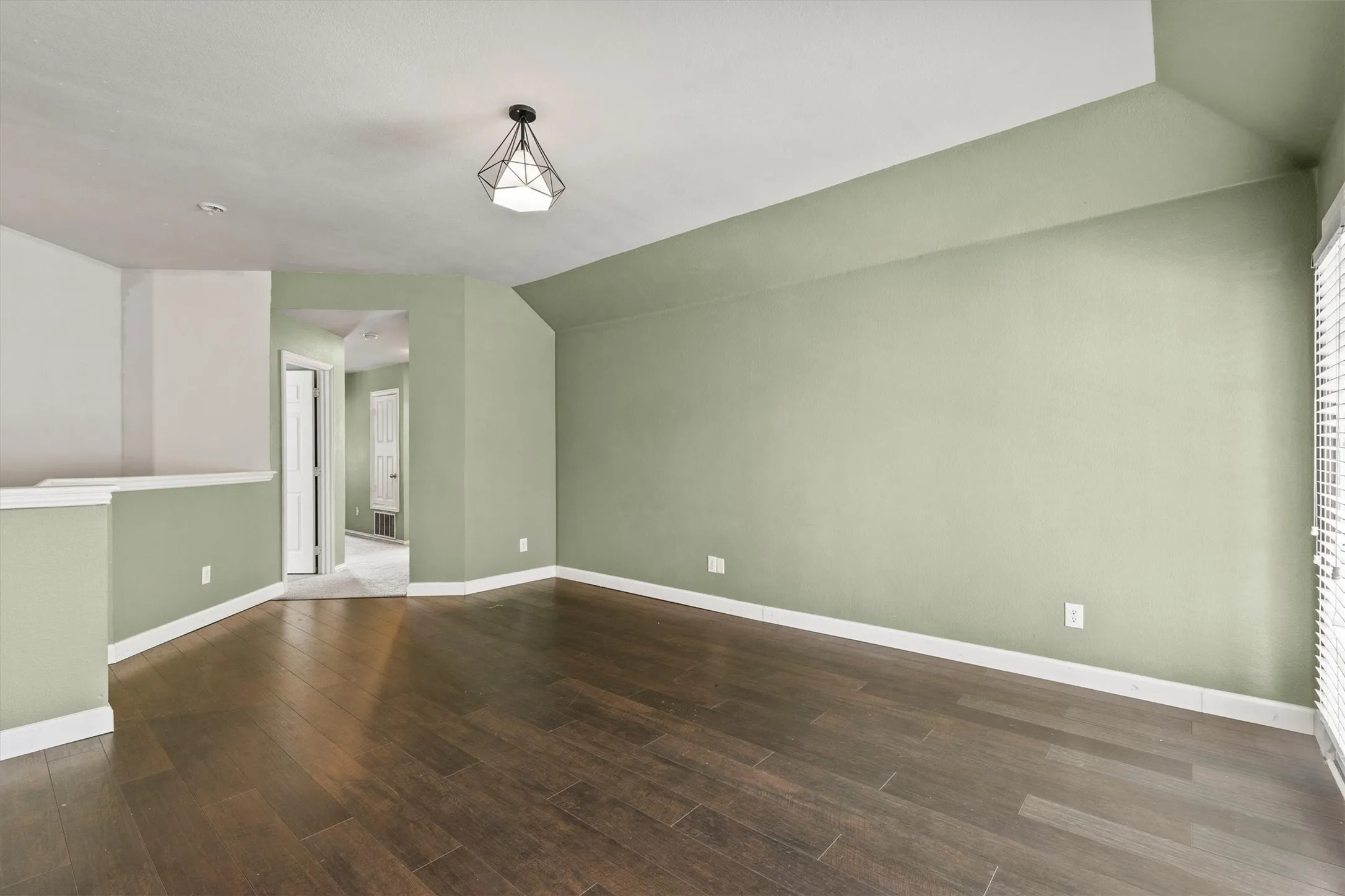 Spacious upper-level room featuring wood-finish flooring, a contemporary geometric light fixture, light green wall paint, white trim, and a window with horizontal blinds