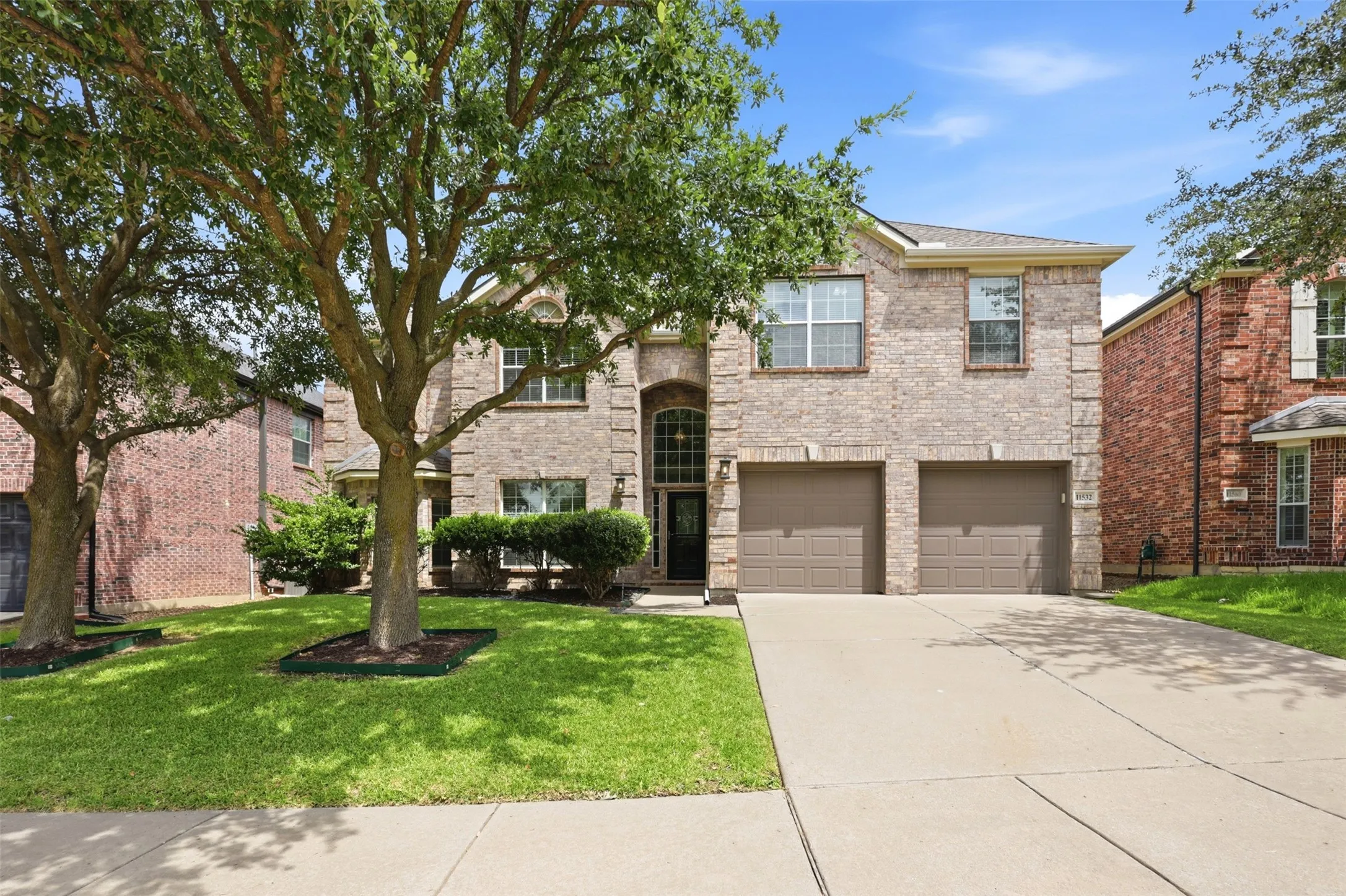 Two-story brick exterior featuring a multi-pane glass entry door, arched portico, dual two-car garage doors, and mature trees in the front yard.