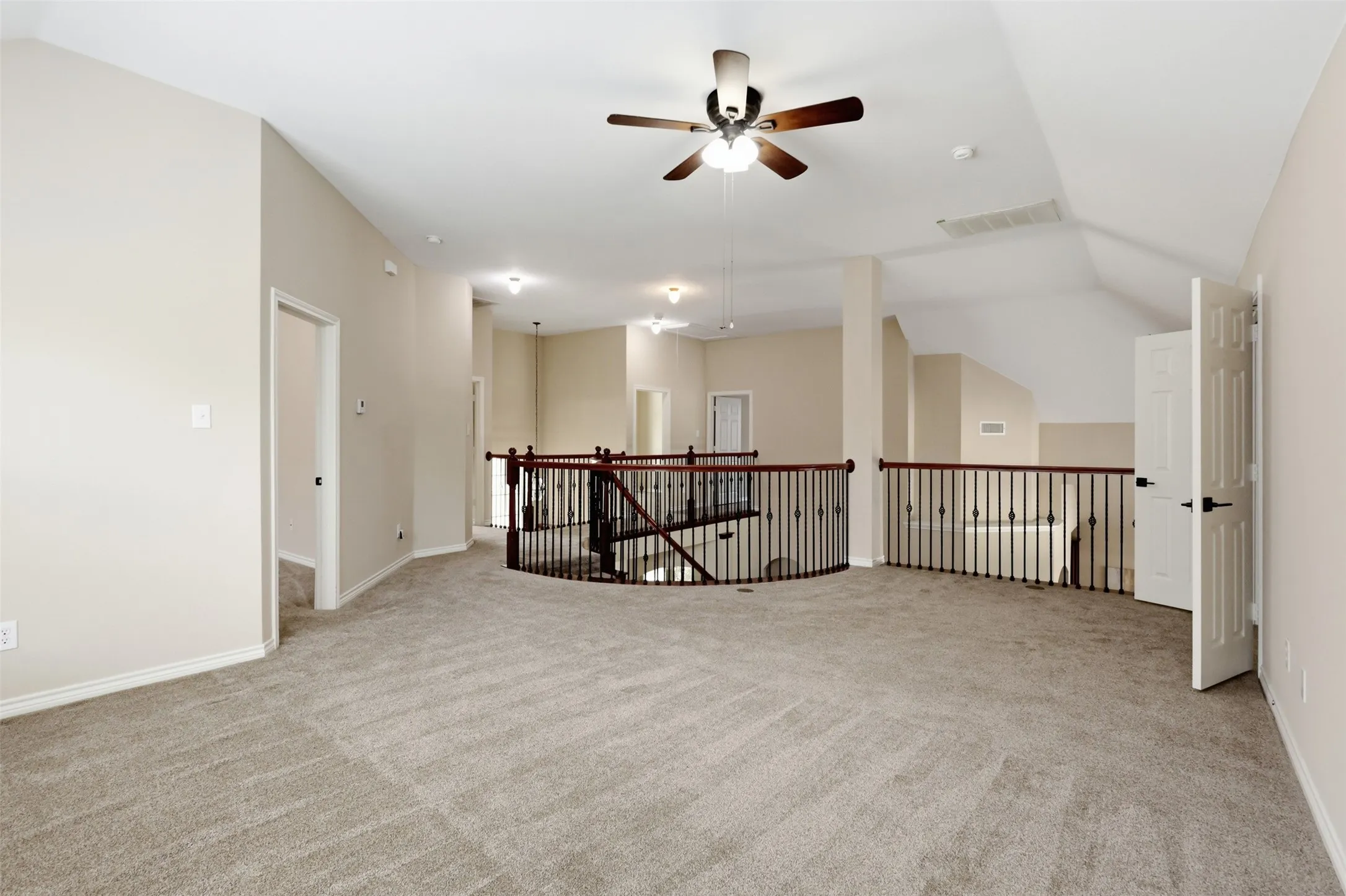 Spacious carpeted upper-level area featuring a decorative metal and wood railing overlooking a lower space, a ceiling fan with integrated lighting, recessed lighting, and neutral wall tones.