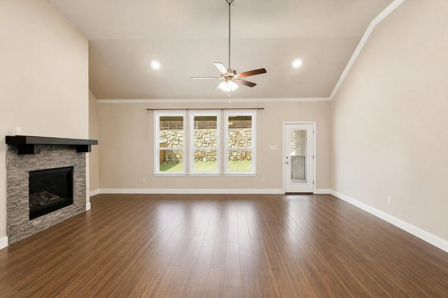Spacious living area featuring wood-finish flooring, a stone-clad fireplace with a dark mantel, and vaulted ceilings with a ceiling fan