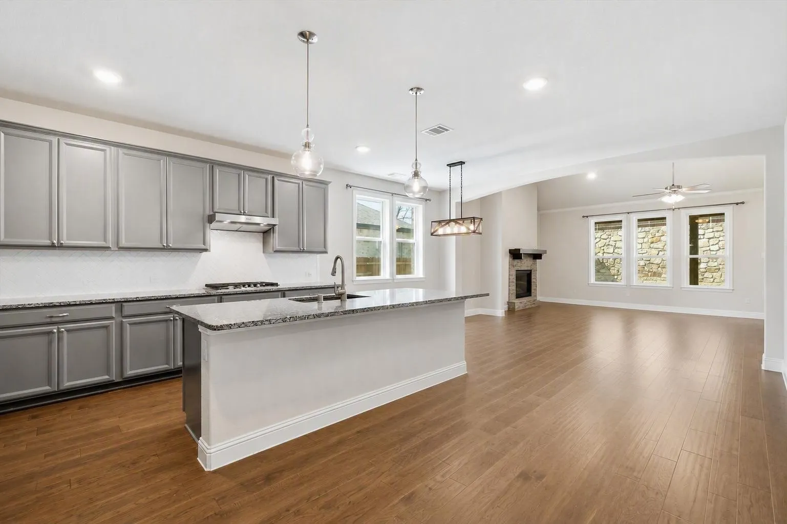 Open-concept living space featuring a kitchen island with sink, gray cabinetry, wood-finish flooring, a stone fireplace, and multiple windows